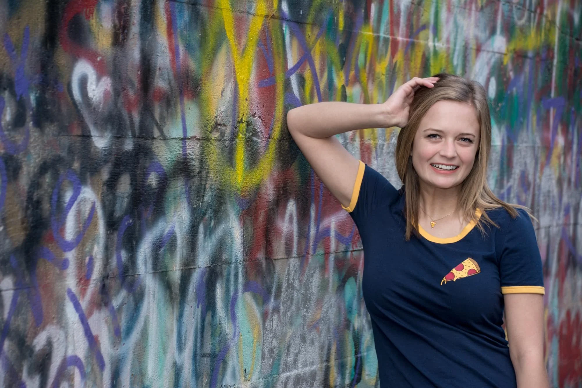 Close-up senior photo of a girl smiling and leaning against a graffiti wall in Columbia IL, wearing a graphic shirt with a slice of pizza on it with her hand on her head.