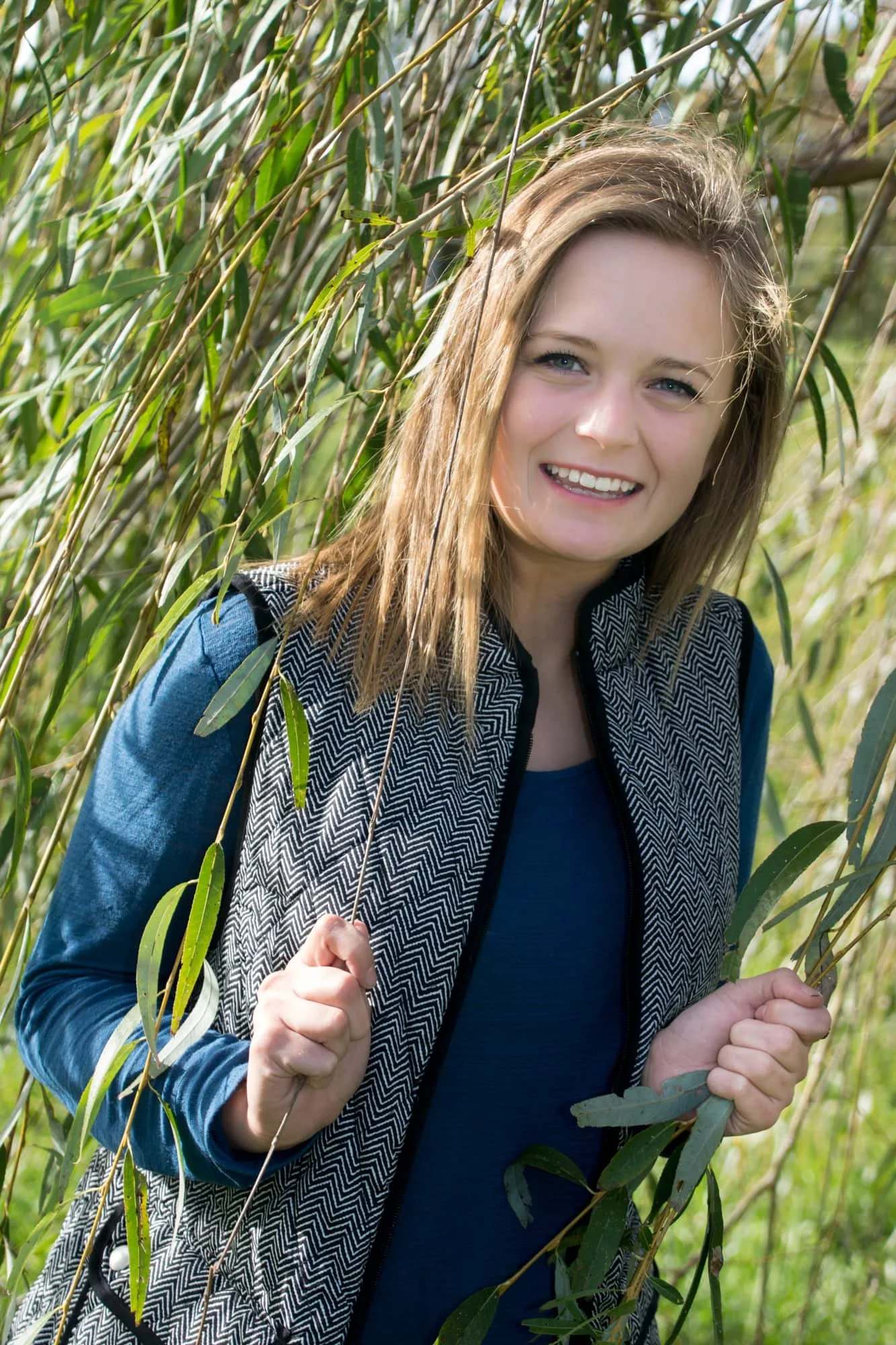 Senior picture of a girl standing inside a willow tree, smiling and grabbing the leaves while wearing a black vest and a teal long-sleeve shirt at Lakeview Park in Waterloo IL.