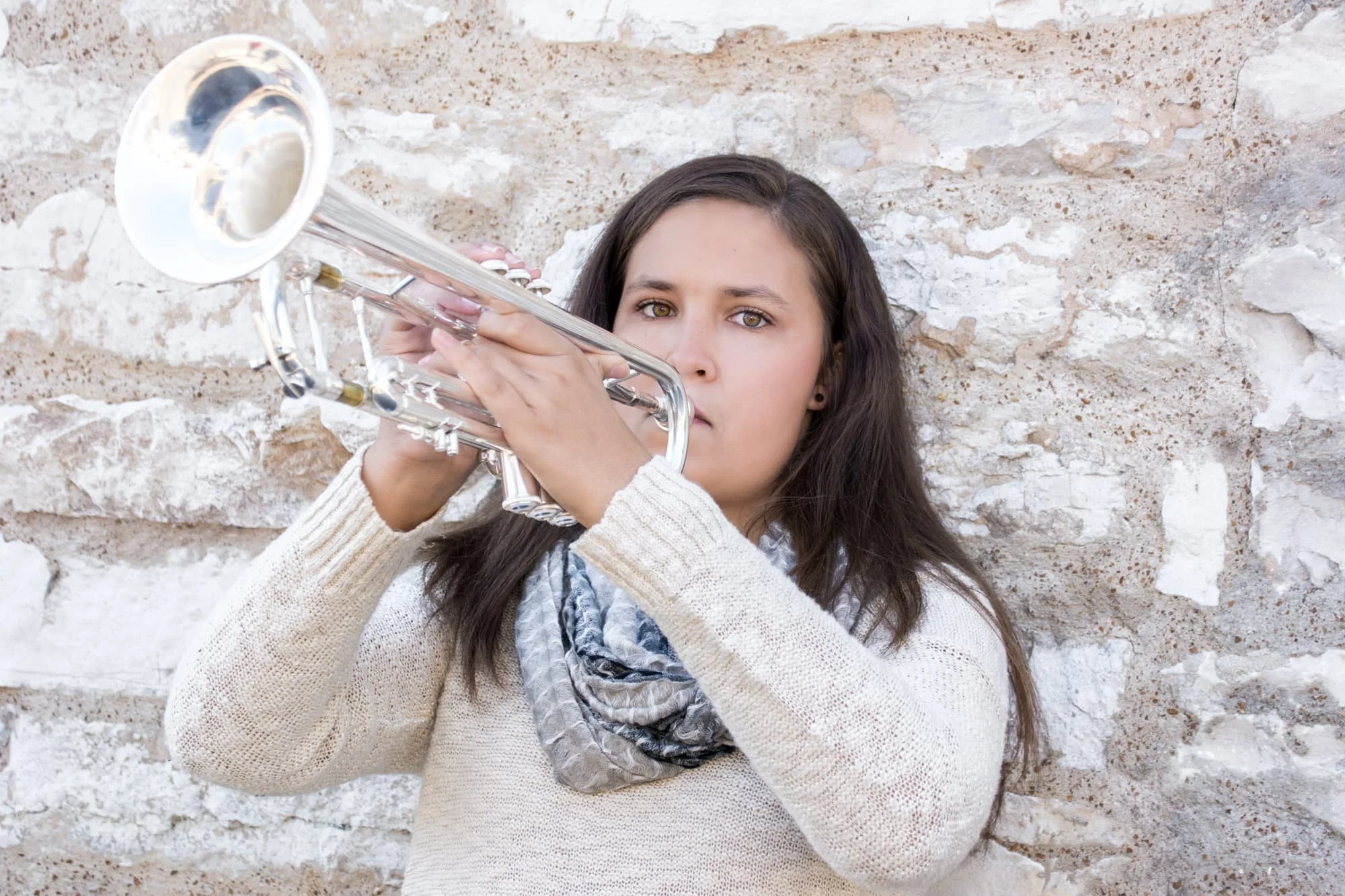 Senior girl playing her trumpet while leaning against an old stone wall, wearing a cream sweater and a tan scarf for a unique, historic-styled senior portrait in Maeystown IL.