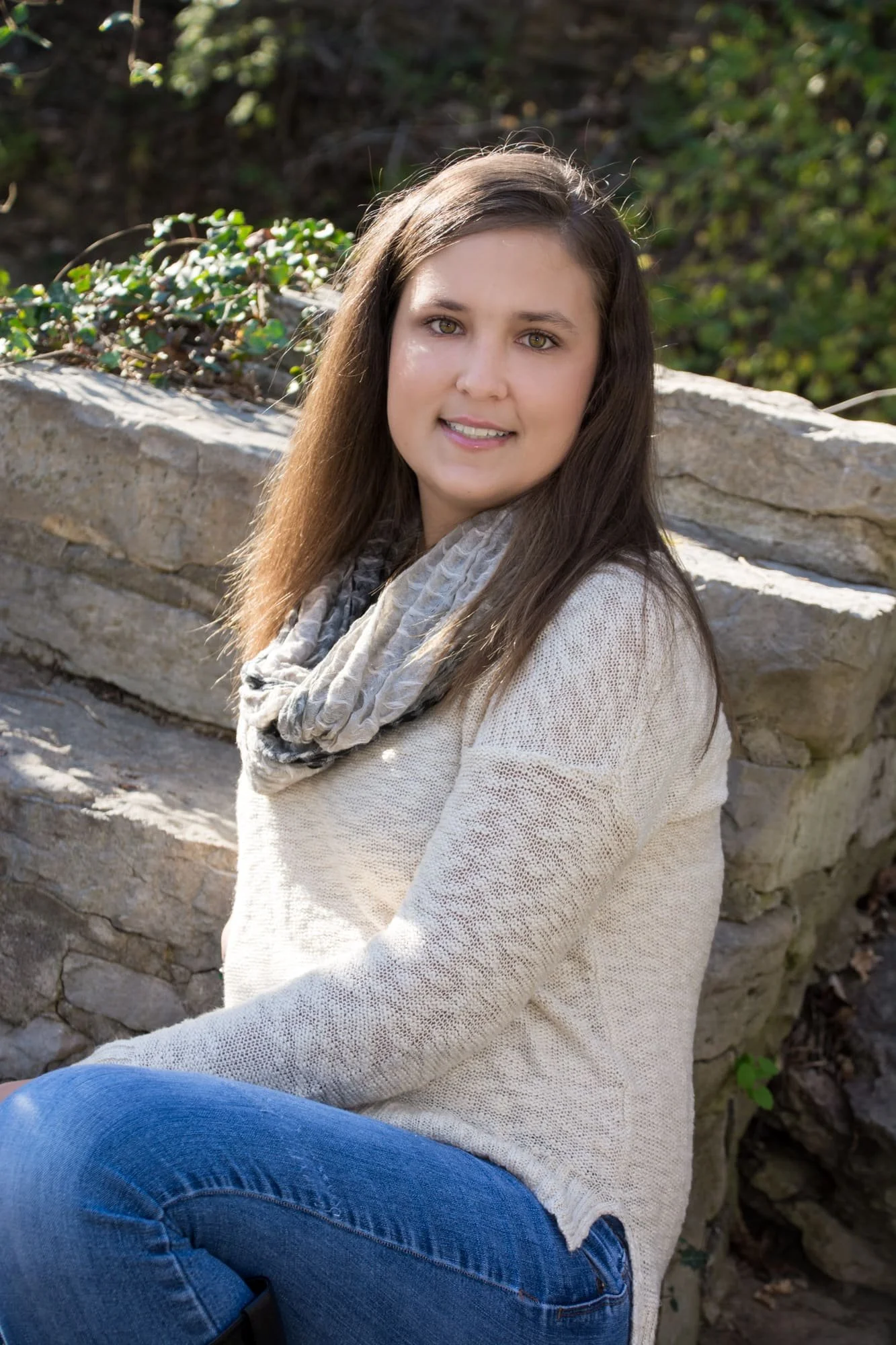 Senior girl wearing a cream sweater, tan scarf, and blue jeans, sitting sideways on stone steps tucked into the woods for a rustic senior picture in Maeystown IL.