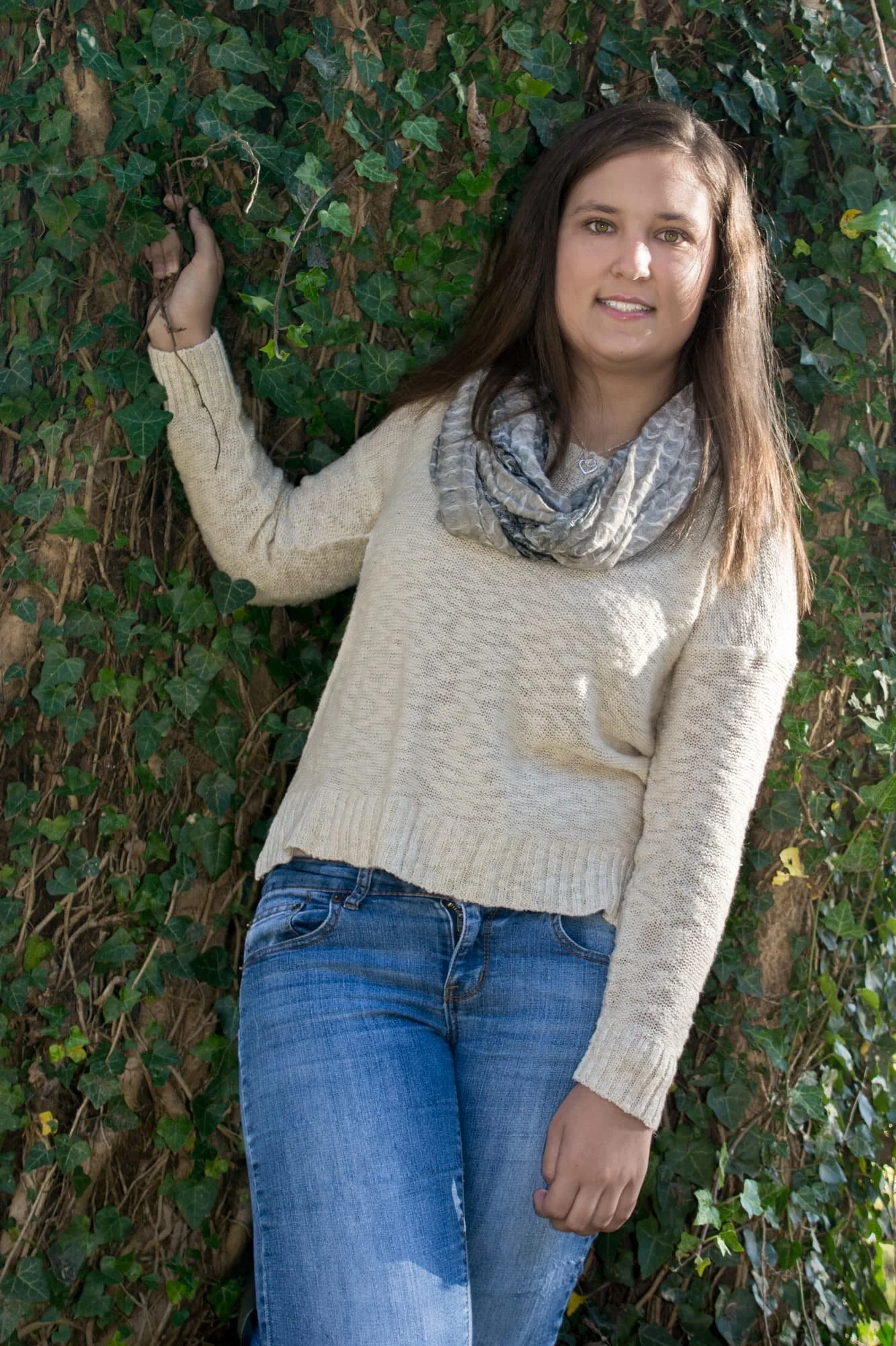Senior photo of a girl standing in front of a tree covered in ivy with her hand resting on the leaves, wearing a cream sweater, tan scarf, and blue jeans in Maeystown IL.