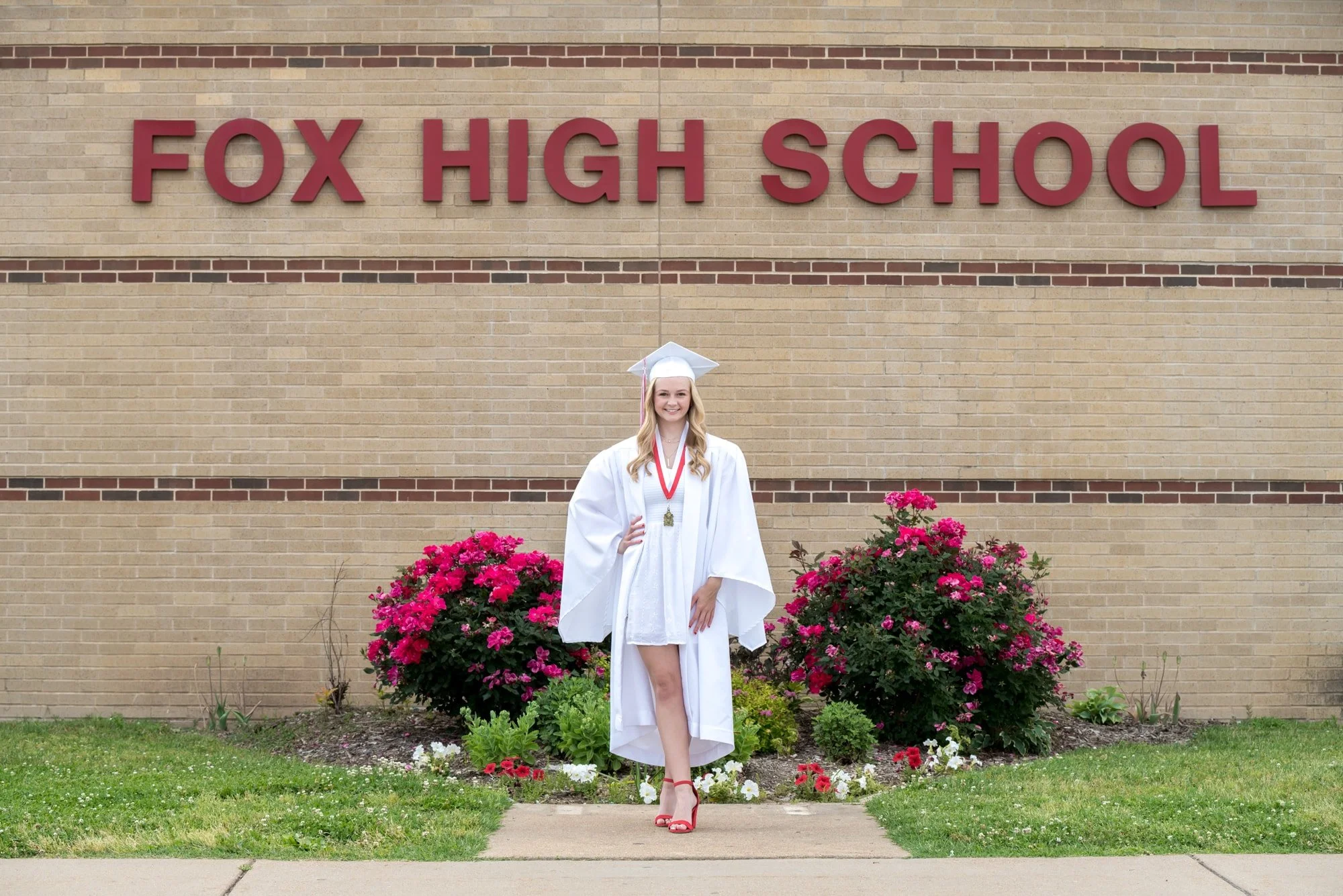 Graduating senior girl smiling in front of the Fox High School sign in Arnold MO, wearing a white graduation cap and gown over a white dress with vibrant red heels.