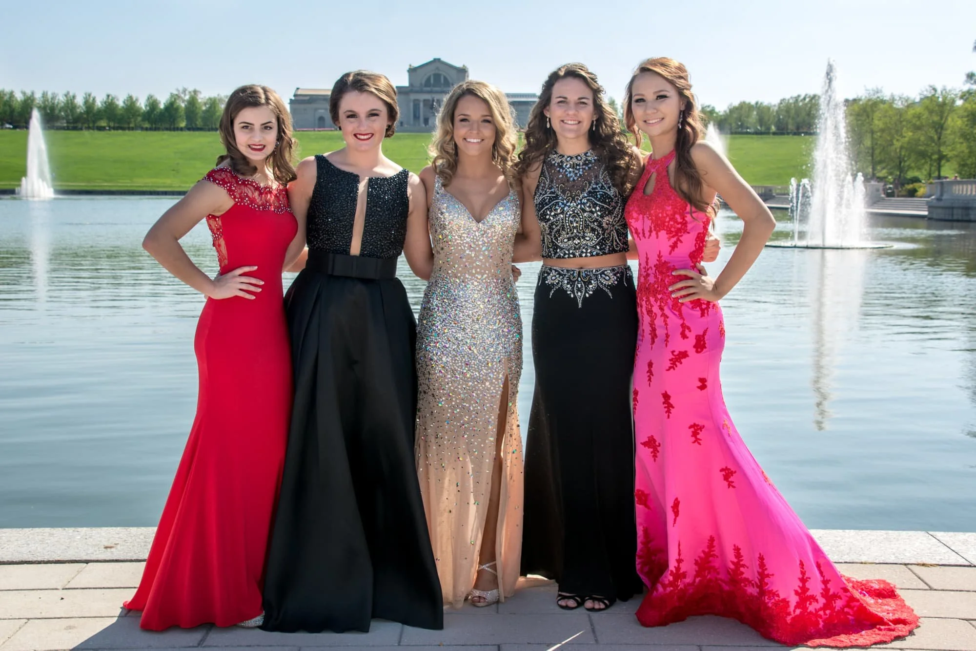 Five girls standing with their arms around each other in front of the Art Hill lake basin at Forest Park, wearing black, pink, red, and tan dresses for a group prom photo.