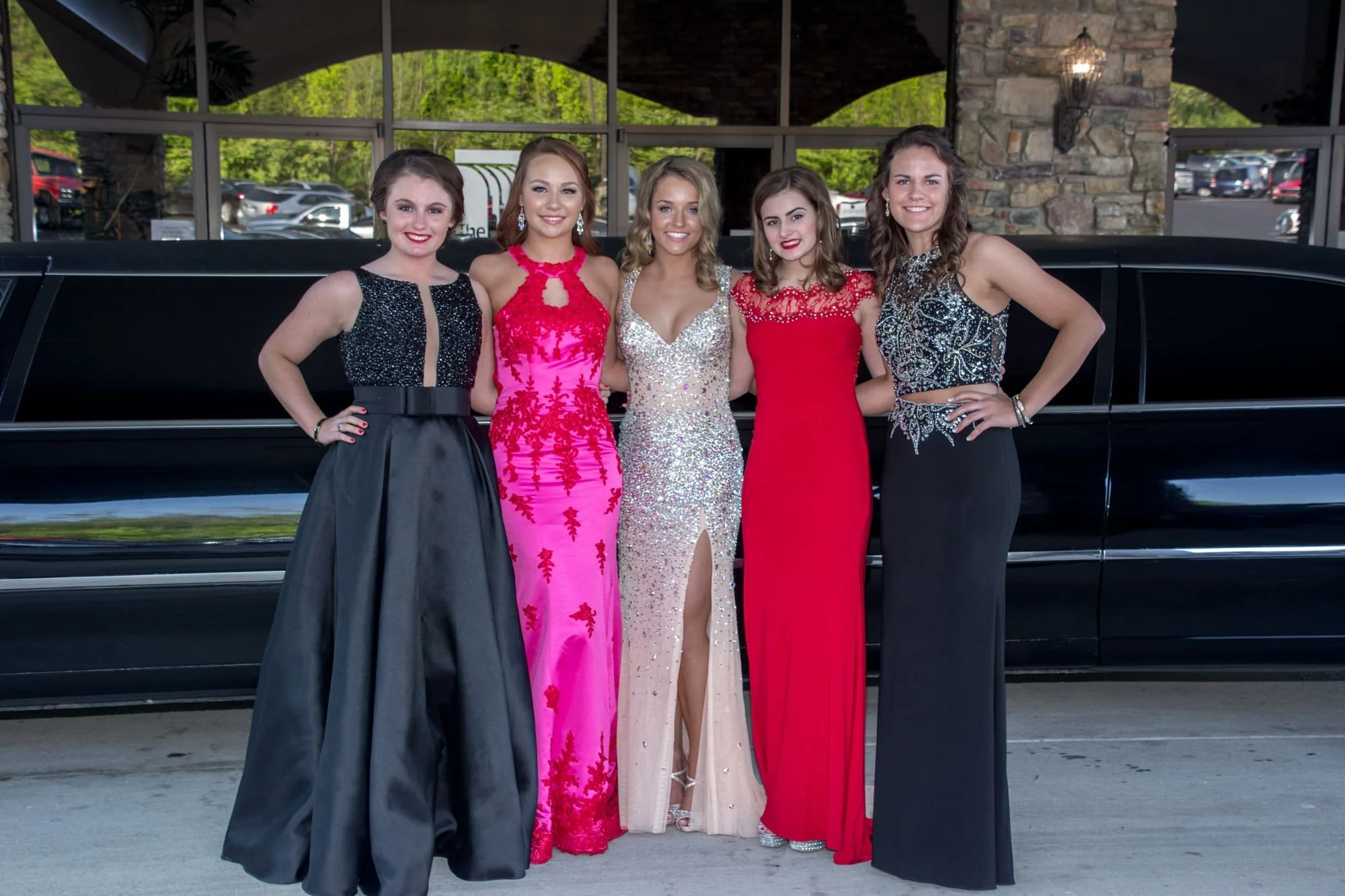 Five senior girls standing with their arms around each other in colorful dresses in front of their limo and venue for prom pictures in Columbia IL.