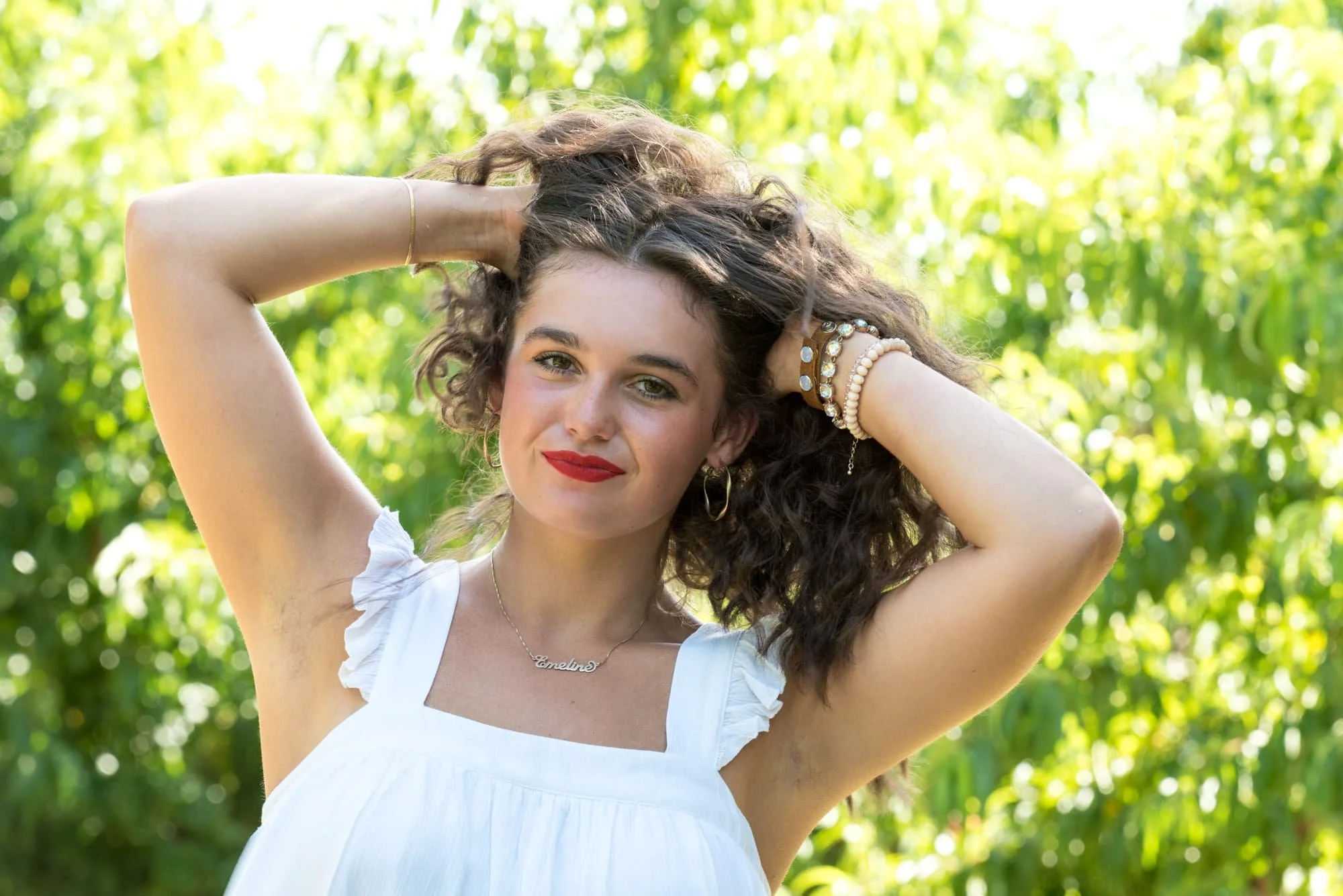 Close-up senior photo of a girl with red lipstick and a soft smile, sitting in front of sun-filled trees with her hands in her hair for a whimsical, sun-drenched portrait at Eckert's.