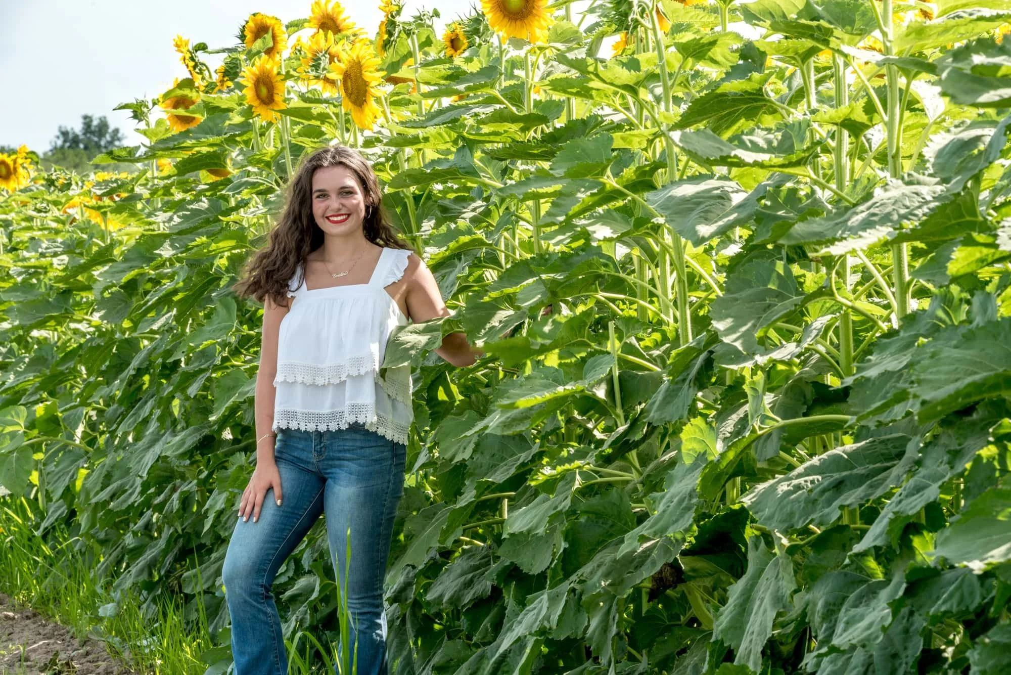 Senior girl wearing a white tank top, blue jeans, and red lipstick, standing alongside a sunflower field with a big smile and her knee bent for a rustic sunflower senior picture at Eckert's.