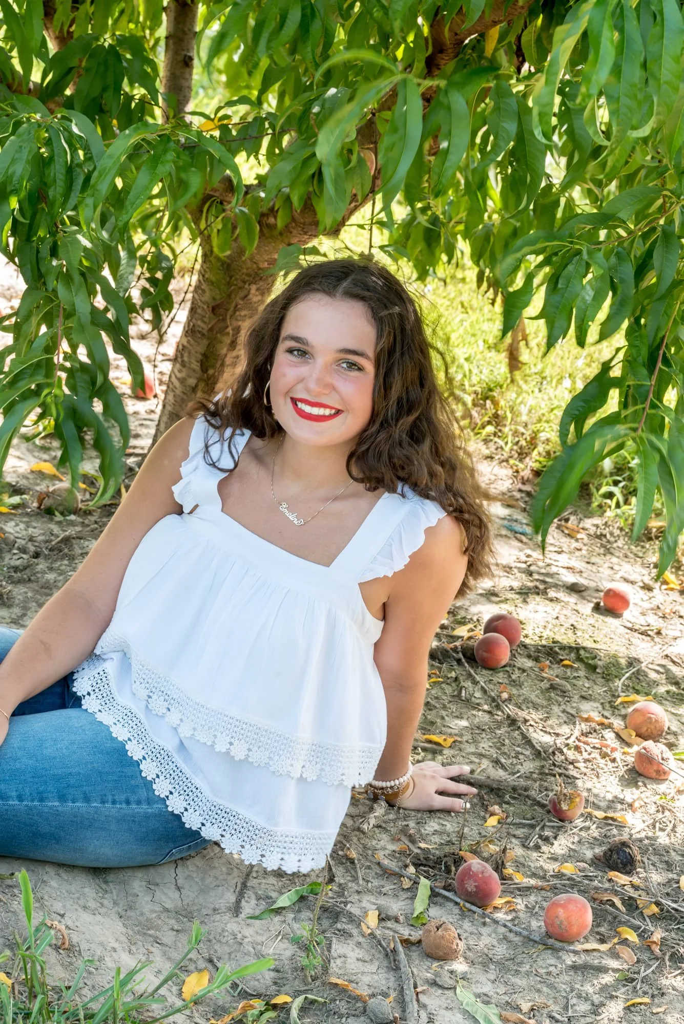 Senior girl laughing and leaning back in the dirt under a peach tree with fresh peaches behind her, wearing a white tank top, blue jeans, and red lipstick for a country-style senior photo at Eckert's.