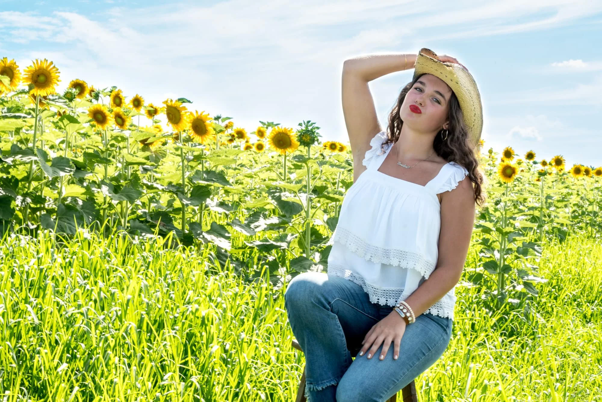 Playful senior picture of a girl making a kissy face while sitting on a stool in a sunny sunflower patch, wearing a white tank, blue jeans, and a cowboy hat at Eckert's Orchard in Belleville IL.