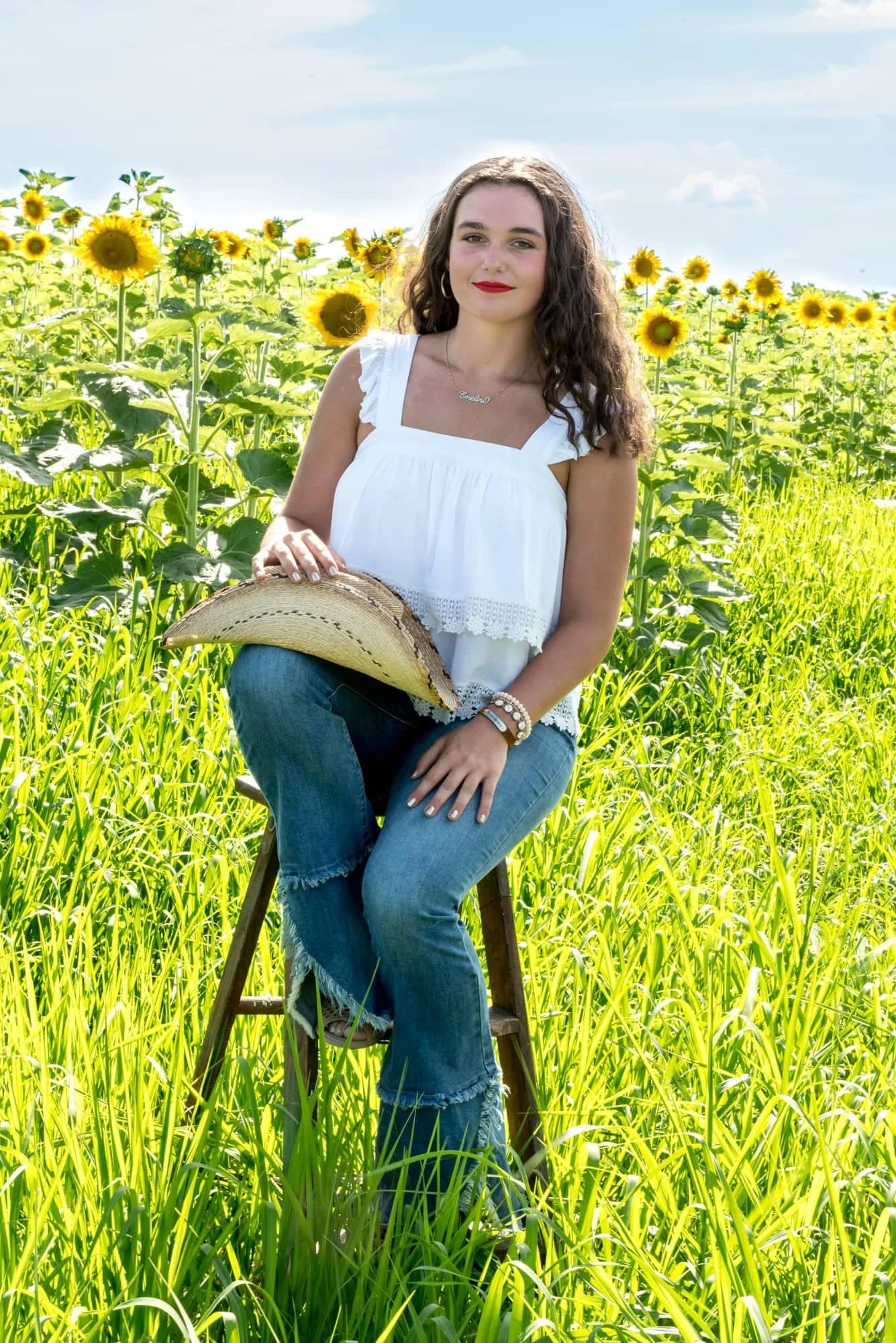 Summer senior portrait by a Belleville IL senior photographer, showing a girl in a white tank top and blue jeans sitting on a stool with a cowboy hat on her lap in front of a sunflower field at Eckert's.