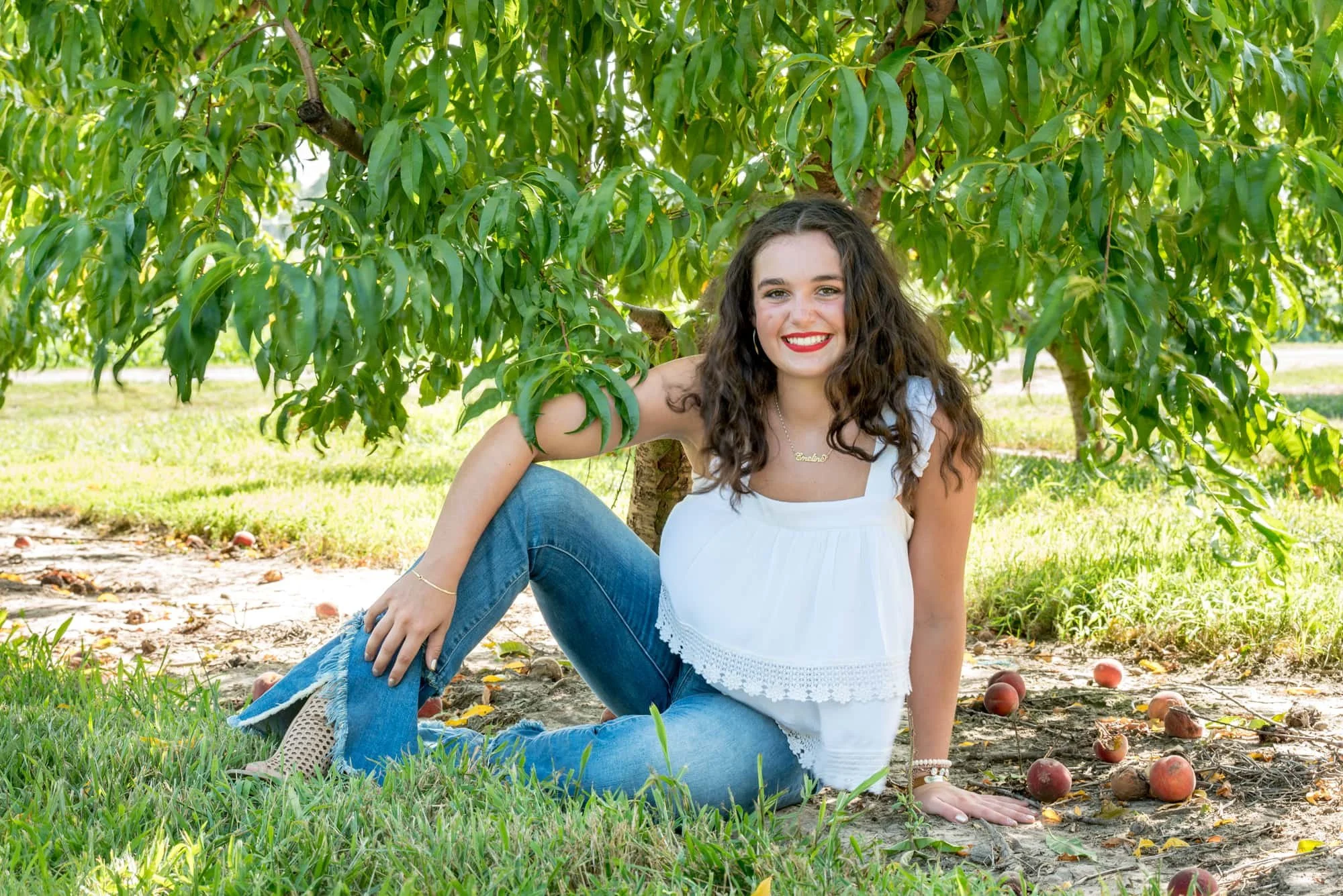 Beautiful country senior photo of a smiling girl wearing tan boots, sitting under a lush peach tree surrounded by fallen fruit with her arms crossed over her leg at an orchard in Belleville, Illinois.