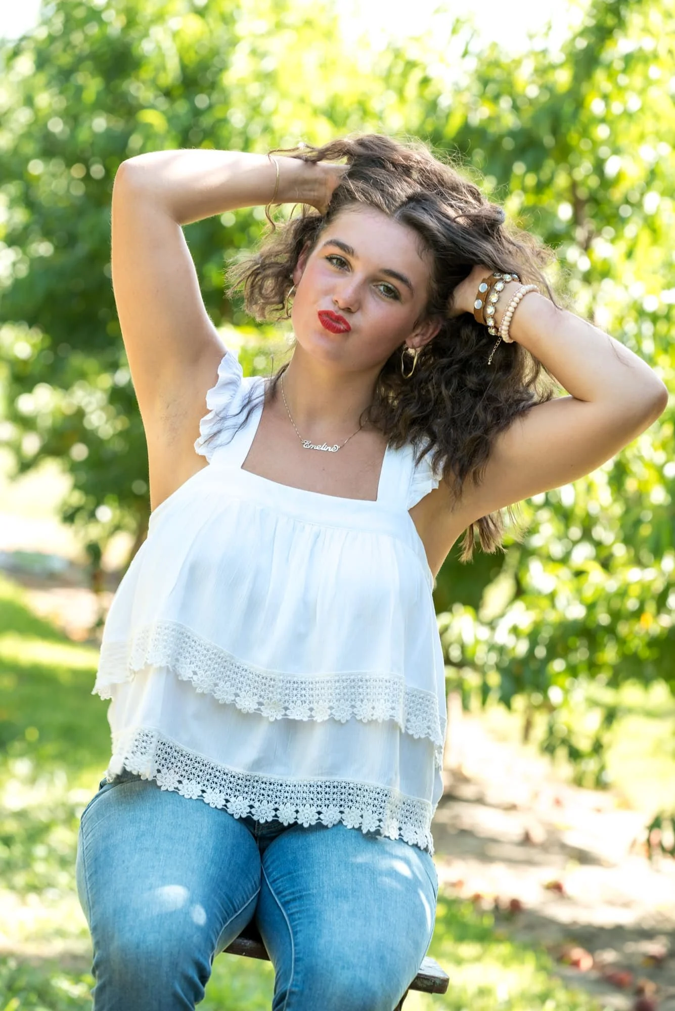 Playful lifestyle senior photo of a girl running her hands through her hair with her head turned sideways, posing on a stool in front of a large tree while blowing a kiss taken by a St. Clair County photographer.