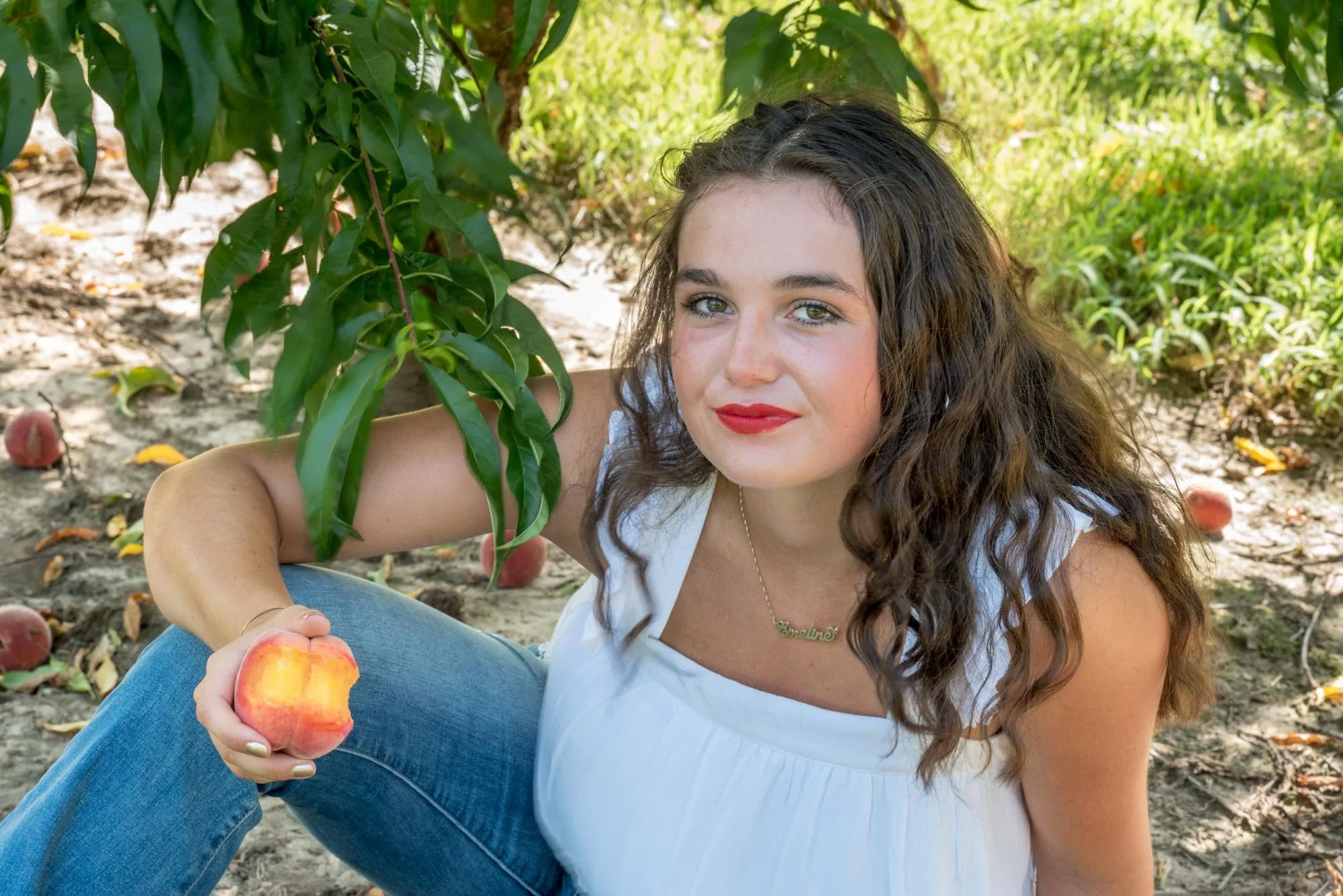 Creative overhead senior photo capturing a girl resting on the ground under a peach tree, holding up a bitten peach with a soft smile as low-hanging branches frame the shot taken by St. Clair County senior photographer.