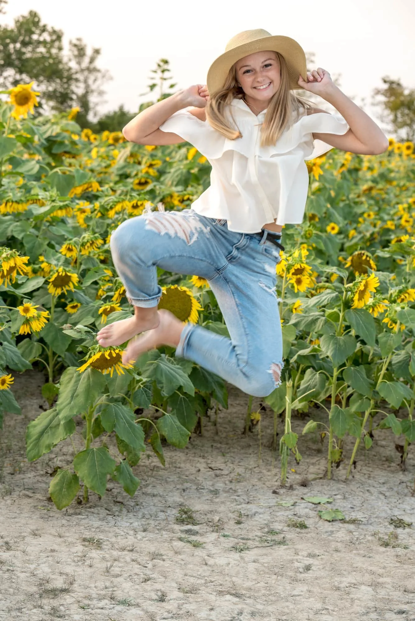 Fun and energetic outdoor senior photo of a barefoot girl in a white top and blue jeans, joyfully clicking her heels together while jumping in the dirt of a sunflower field.