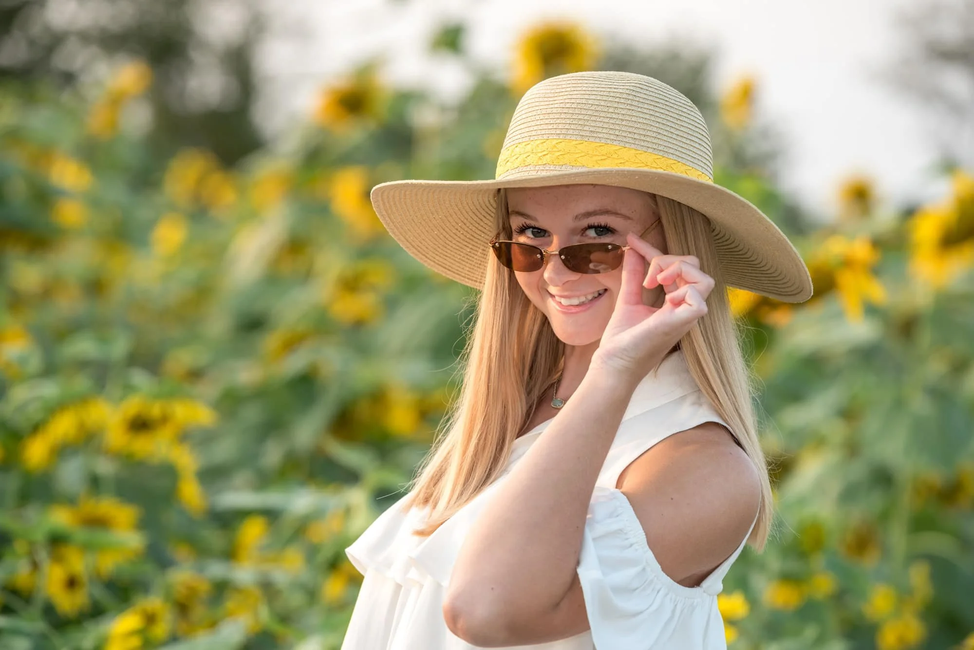 Stylish close-up senior picture featuring a girl wearing a brown floppy hat and sunglasses, smiling with her hand on her shades in front of a wall of sunflowers in Eureka, MO.