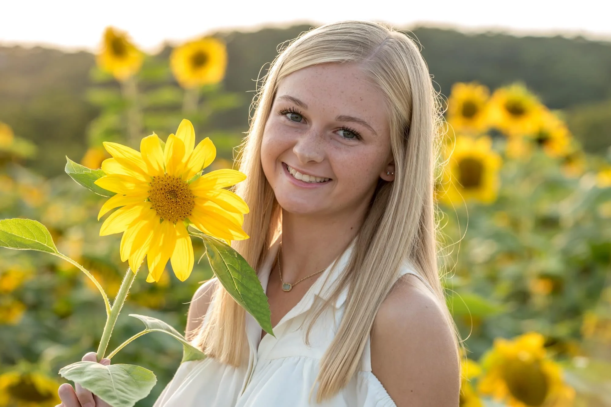 Close-up senior portrait by a Eureka MO senior photographer, featuring a girl in a white tank top smiling and holding a bloom by her face during a vibrant sunset at Brookdale Farms.