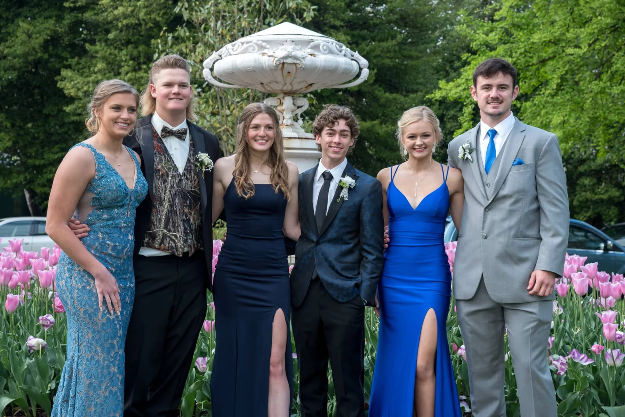 Group of prom-goers in blue dresses and sharp camo, navy, and grey tuxes, posing with their arms around each other in front of blooming pink tulips during a spring prom session at Tower Grove Park.