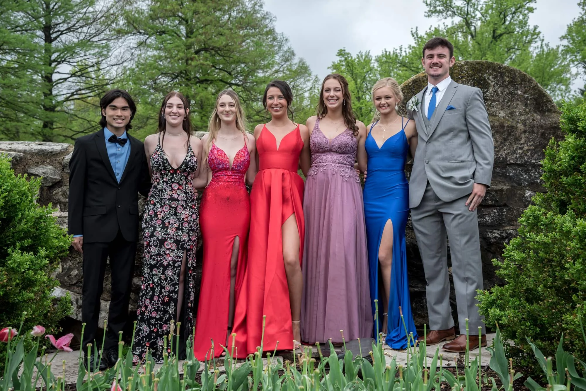 Fun group prom picture capturing a group of friends and couples posing together in front of the rustic stonework and lush spring greenery at Tower Grove Park.