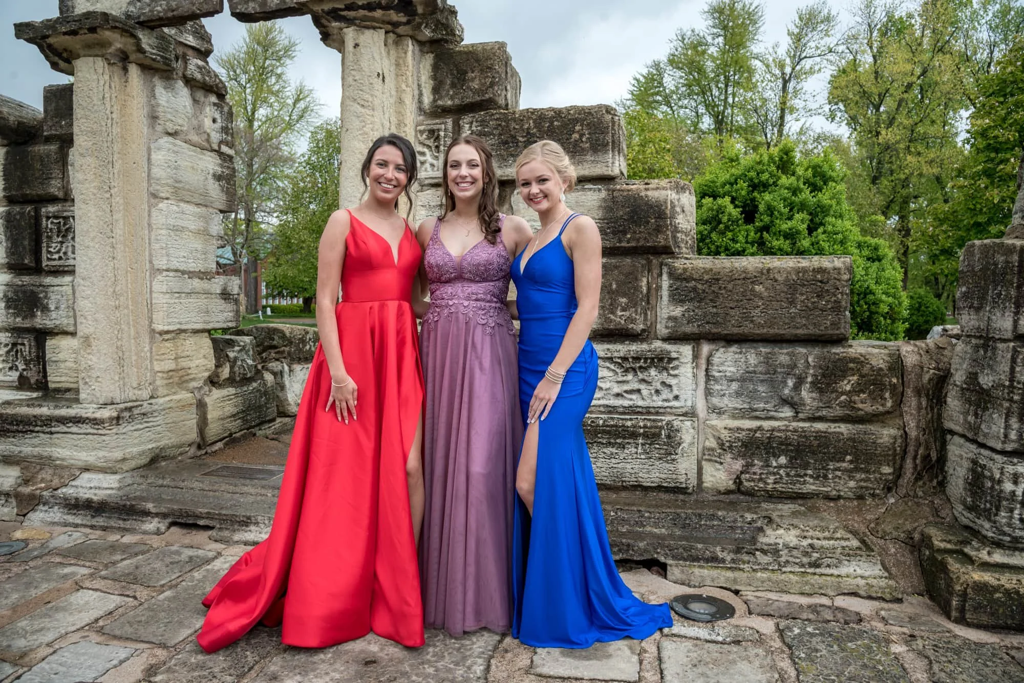 Joyful group prom photo of three friends wearing vibrant red, purple, and royal blue prom dresses, smiling with their arms wrapped around each other in front of a historic stone arch at Tower Grove Park.