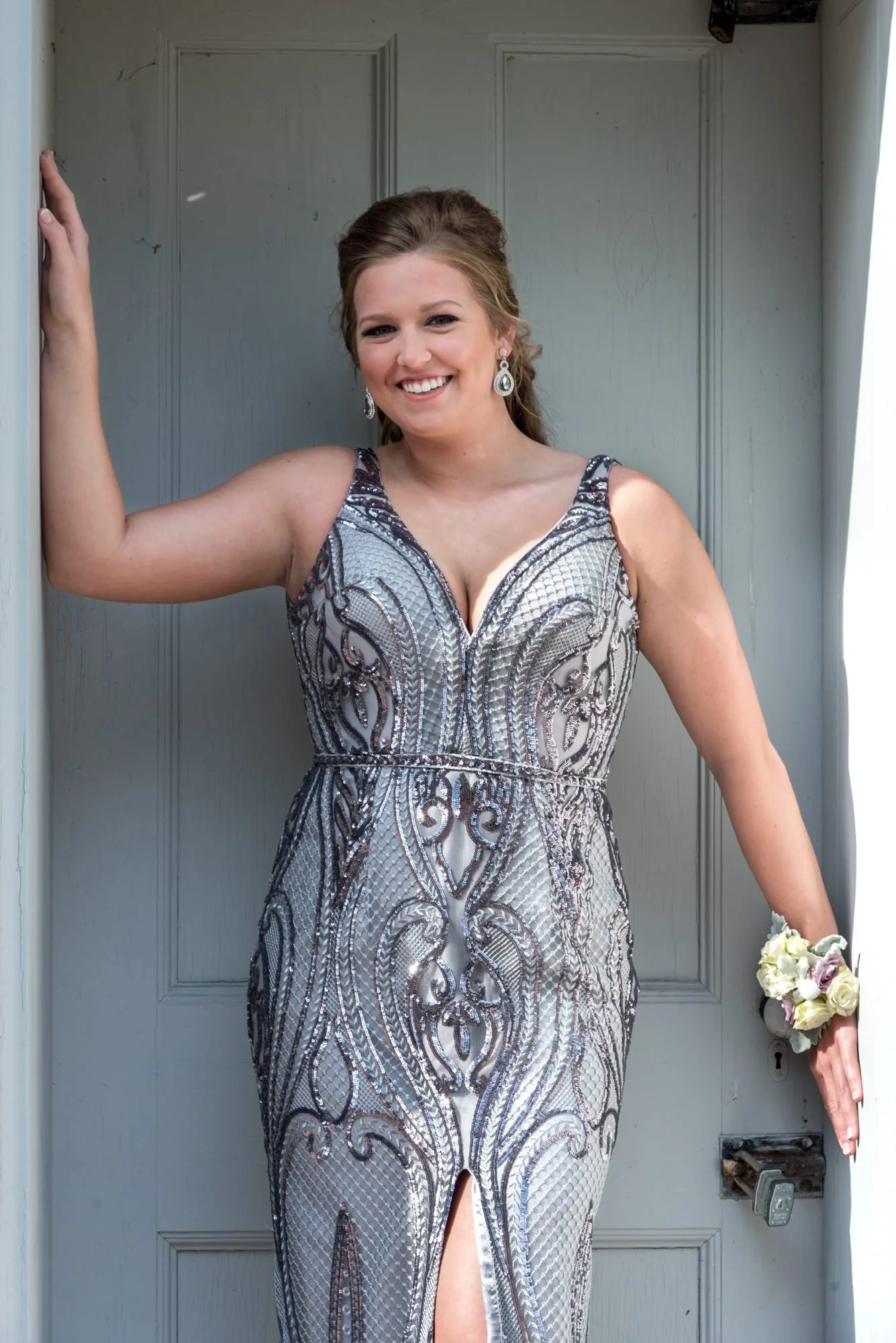 Senior girl wearing a sparkling silver prom dress, standing inside a historic blue doorway with her hands resting on the doorframe for a portrait at Jefferson Barracks Park.