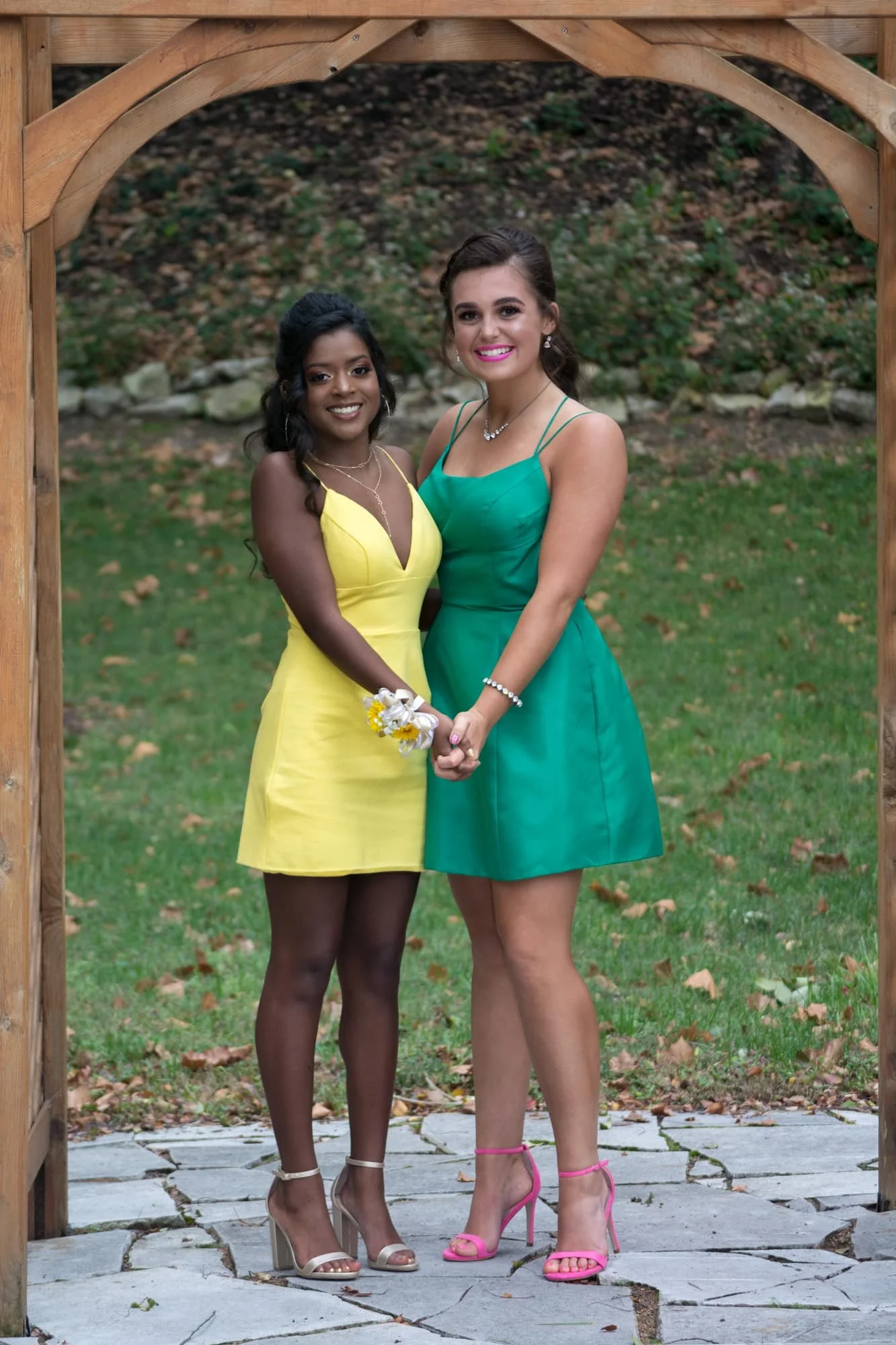High school friends in their yellow and green homecoming dresses standing under a rustic wooden archway, wrapping their arms around each other and holding hands.