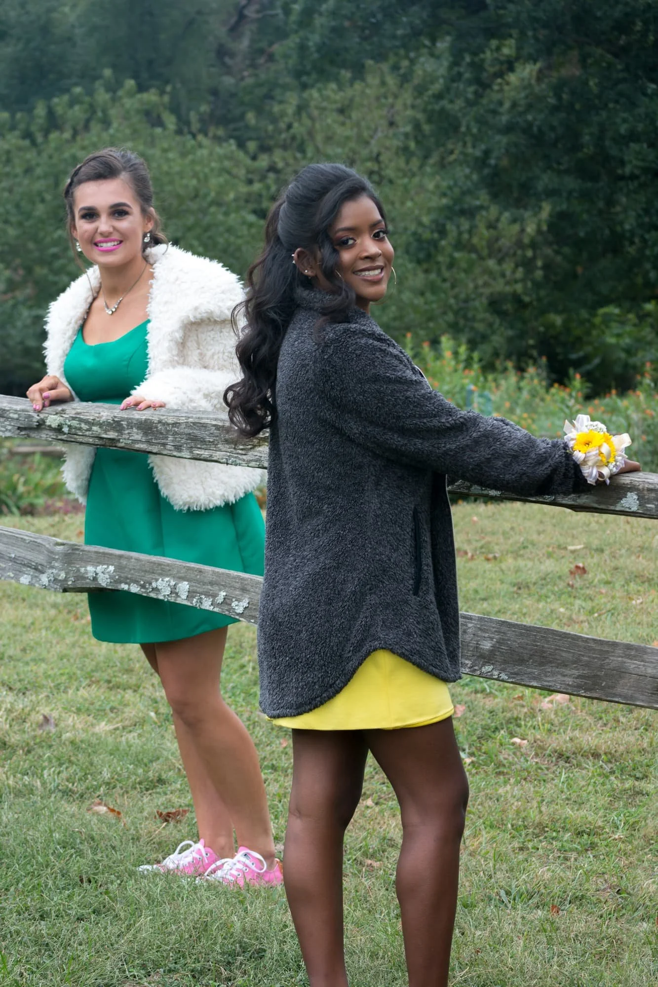 Two homecoming friends wearing elegant yellow and green dresses, leaning on a rustic wooden fence together with a beautiful wooded background at Jefferson Barracks Park.