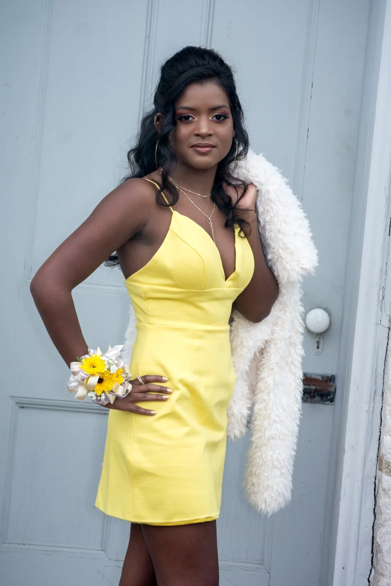 High school homecoming girl posing in a blue doorway at Jefferson Barracks, wearing a yellow formal dress with a cozy fuzzy coat draped over her shoulder.