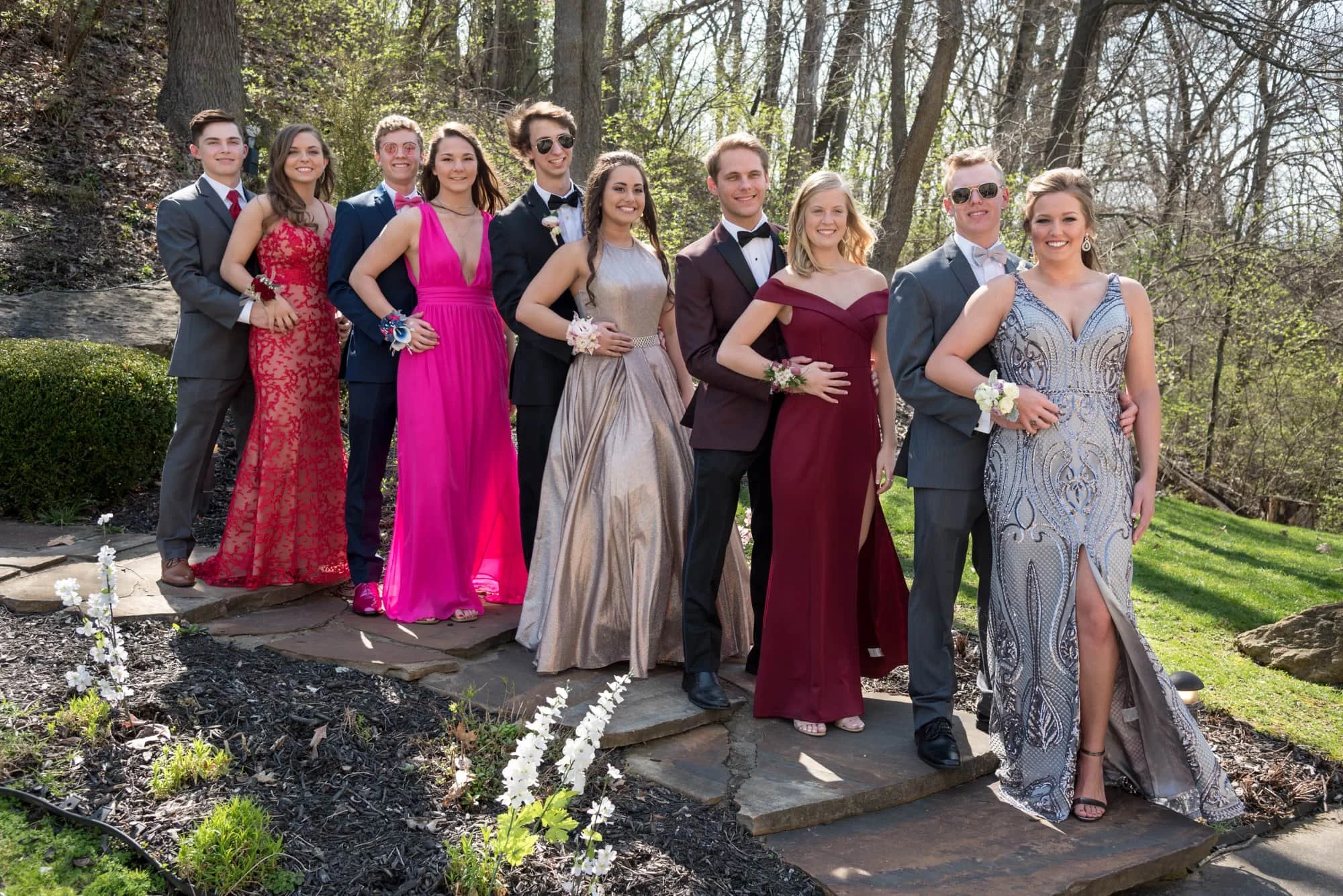 Large prom party of couples posing together with their dates on a rustic stone pathway in the woods, with the girls wearing beautiful silver, red, and pink dresses for a formal group prom photo.