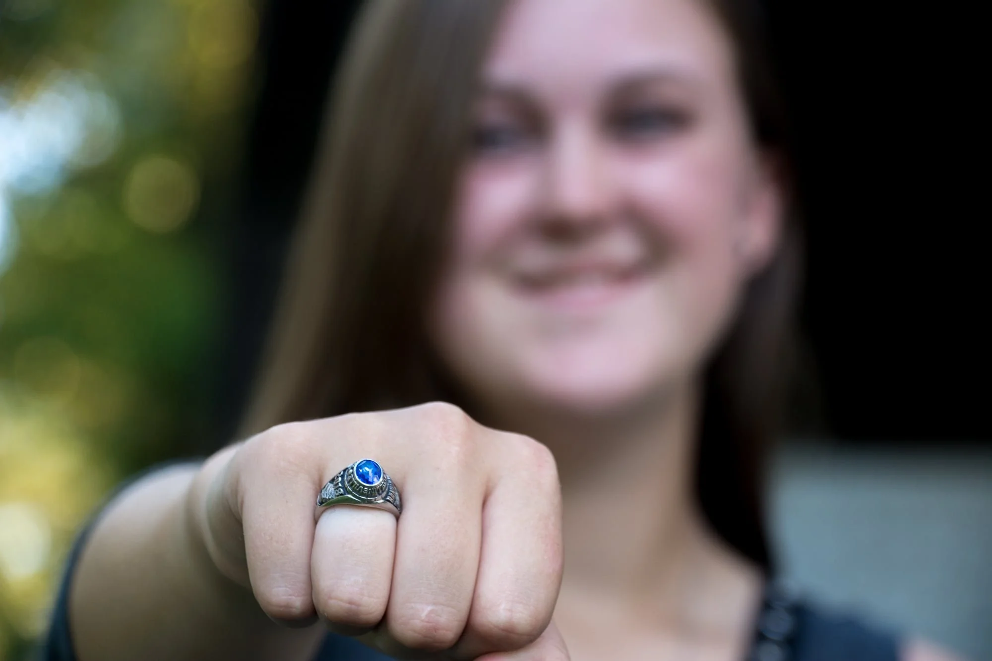 Creative close-up senior picture taken by a St. Louis senior photographer, featuring a sharp focus on a girl's high school class ring on her hand with a beautiful, softly blurred park background.