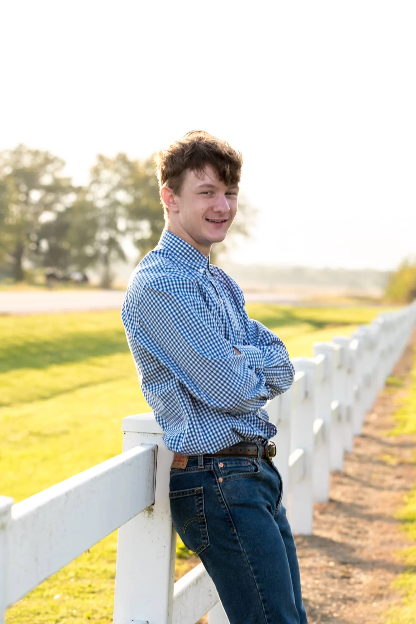 Stunning sunset senior picture capturing a boy in a plaid button-up shirt and blue jeans, standing with his arms crossed while leaning against a clean white fence taken by a Freeburg, IL photographer.