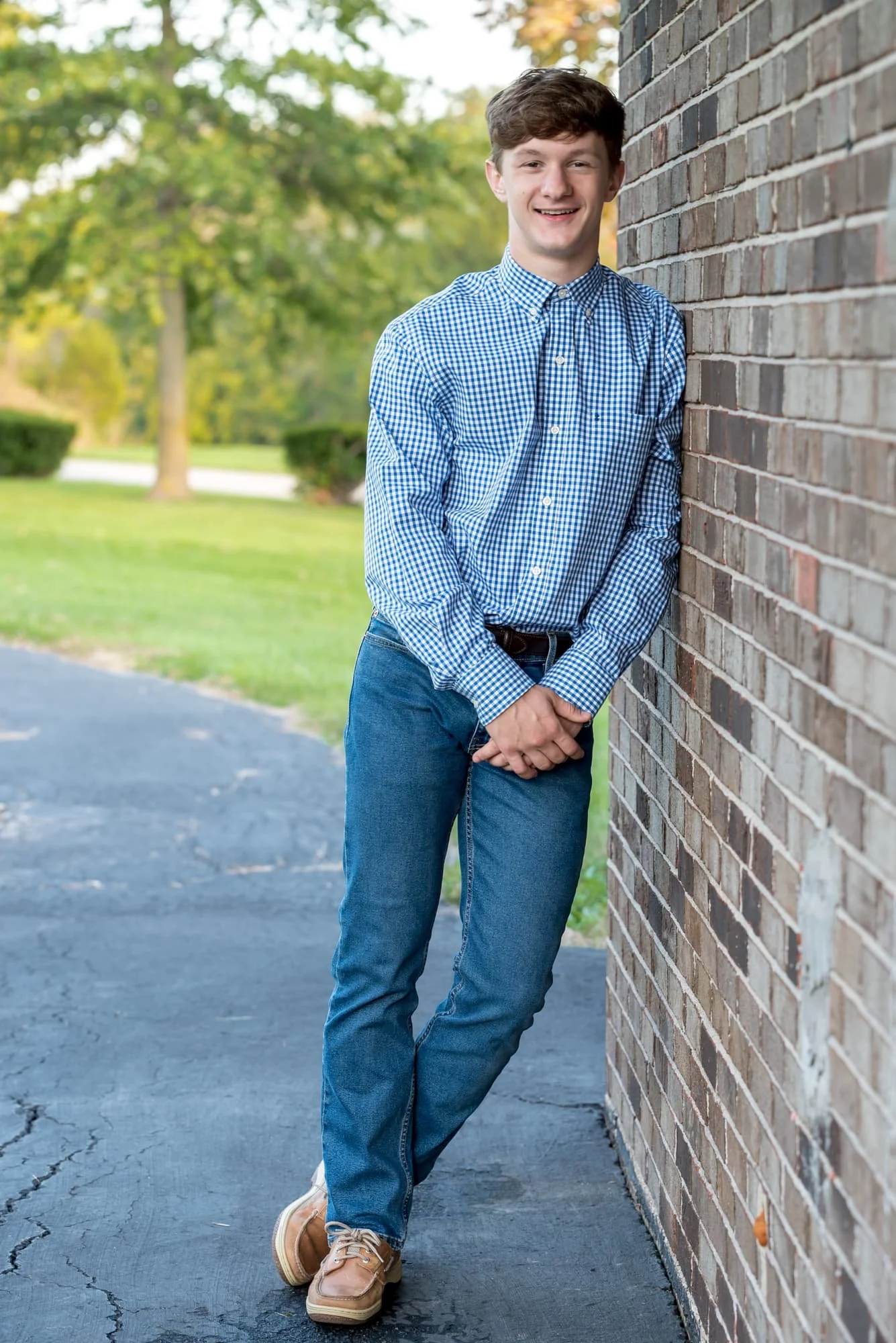 Cheerful autumn senior photo of a smiling boy wearing a blue and white button-up shirt, blue jeans, and brown shoes, clasping his hands together while leaning against a brick building with fall foliage behind him in Freeburg, IL.
