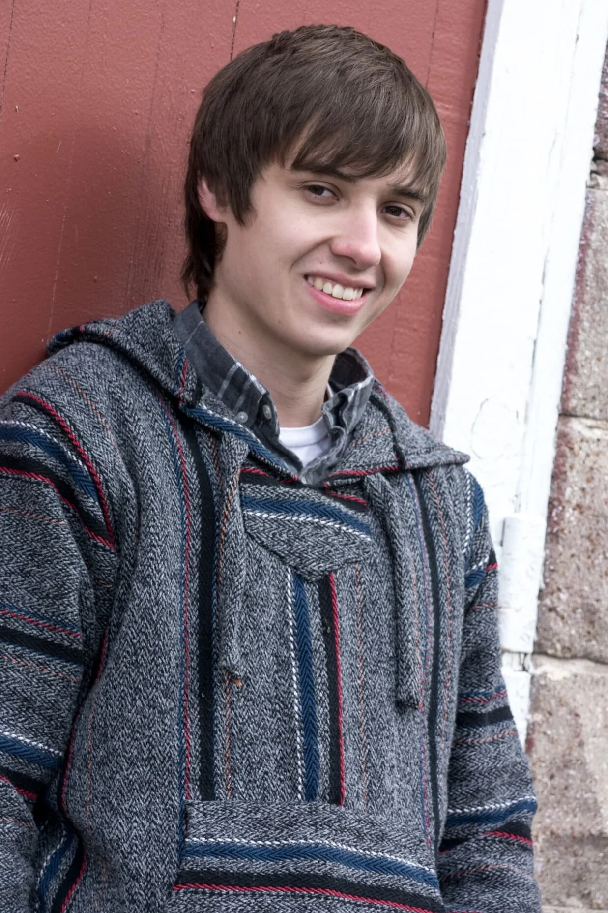 Senior guy wearing a grey sweater, standing in front of a red barn door during his senior photo session at Bee Tree Park.