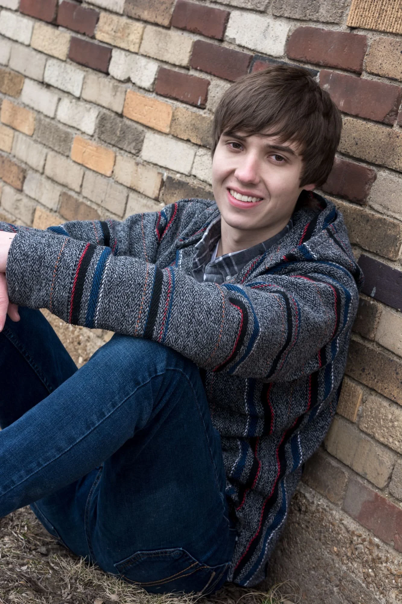 Senior photo of a guy sitting on the ground and leaning against a historic brick wall with his arms resting across his knees at Bee Tree Park in Oakville.