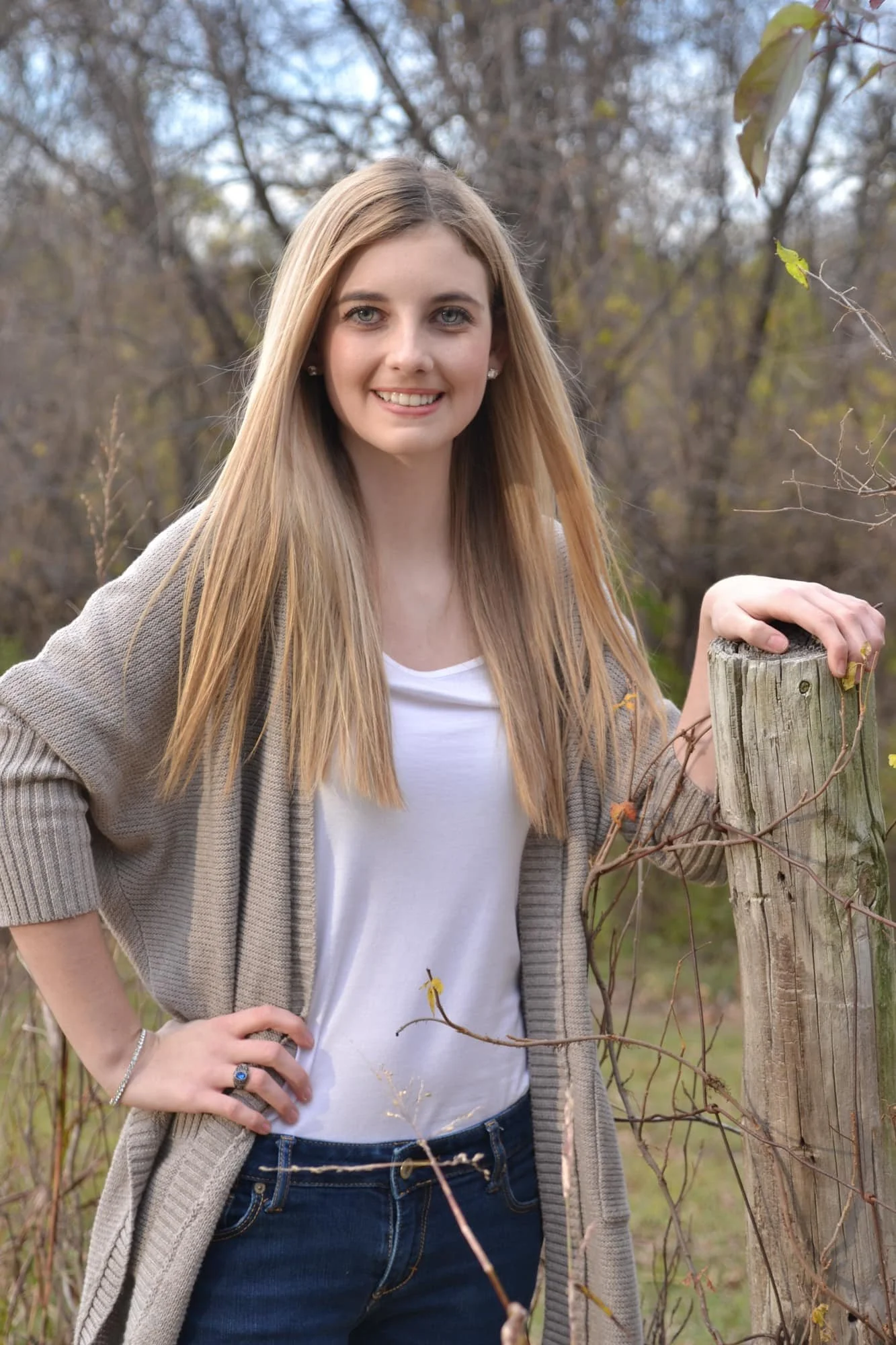 Senior picture of a girl with straight blonde hair blowing in the wind, wearing a tan sweater over a white shirt and blue jeans while standing next to a fence with her hand on a wooden post at Bee Tree Park.