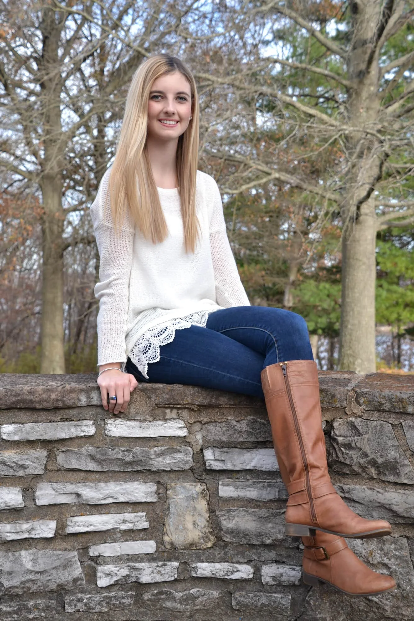 Senior girl wearing a white long-sleeve shirt, blue jeans, and brown boots, sitting on a stone wall during fall with her legs crossed for her senior picture at Bee Tree Park.