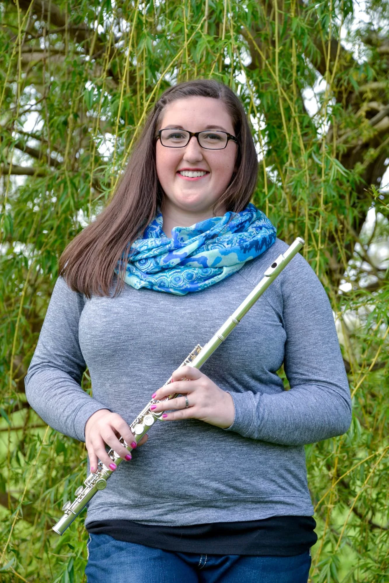 Senior girl wearing glasses, a blue scarf, and a gray shirt, standing in front of a willow tree while holding her flute and smiling for her senior photos at Bolm-Schuhkraft Park.