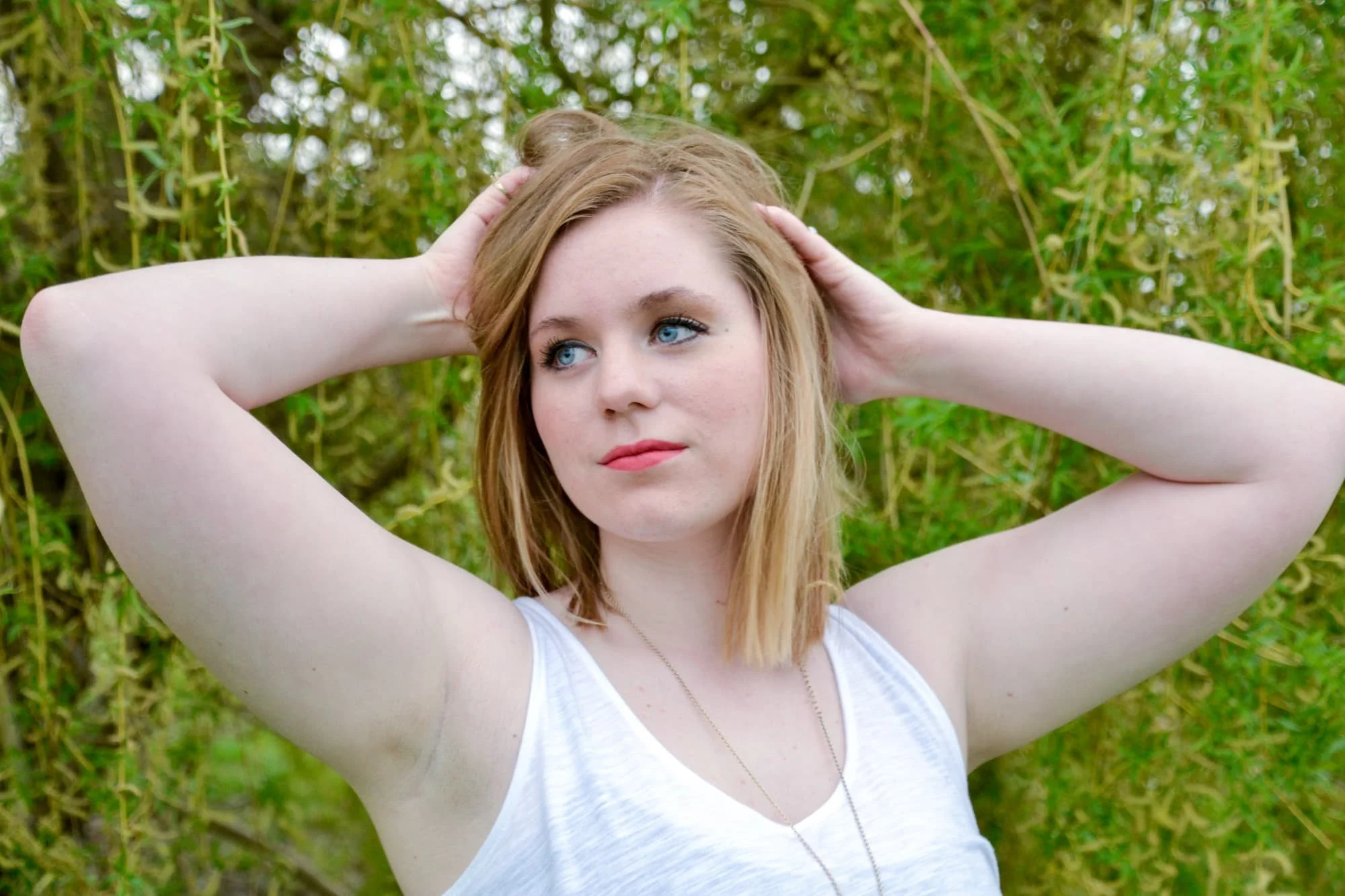 Senior picture of a girl standing in front of a willow tree at Bolm-Schuhkraft Park with her hands in her hair while looking off to the side, wearing a white tank top.