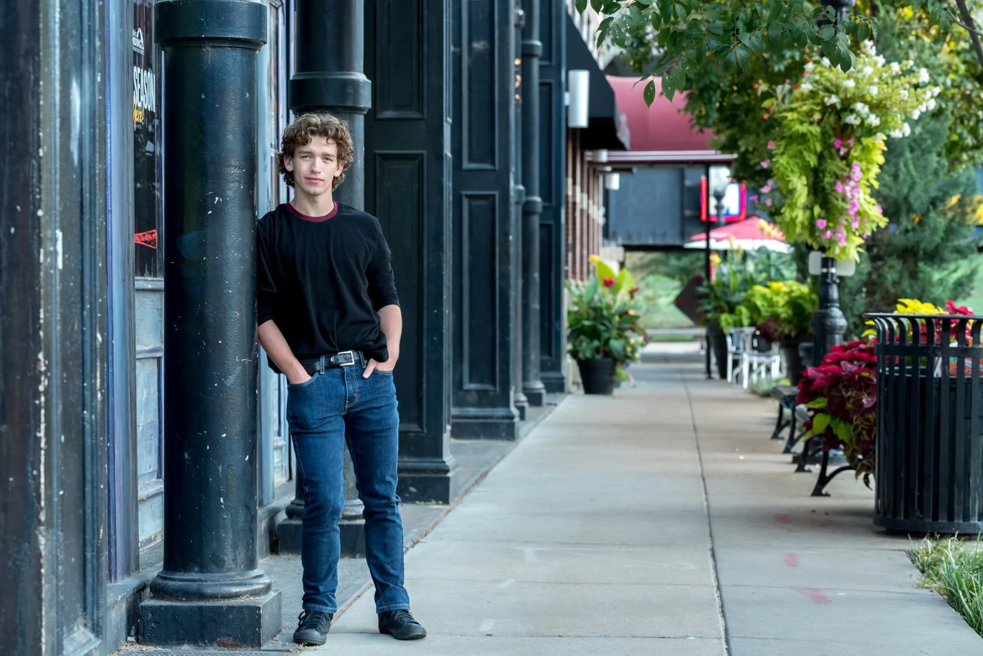 Serious downtown senior photo of a boy wearing a black sweater, blue jeans, and black tennis shoes, standing with his hands in his pockets while leaning against a dark metal beam at Laclede's Landing in St. Louis.