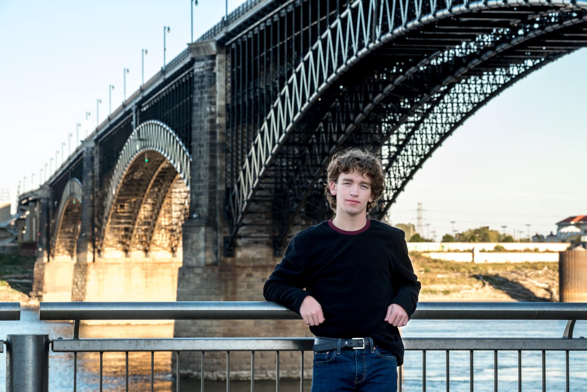 Classic St. Louis senior picture featuring a boy in a black sweater and blue jeans, leaning back against a riverfront railing with the Mississippi River and historic Eads Bridge in the background.