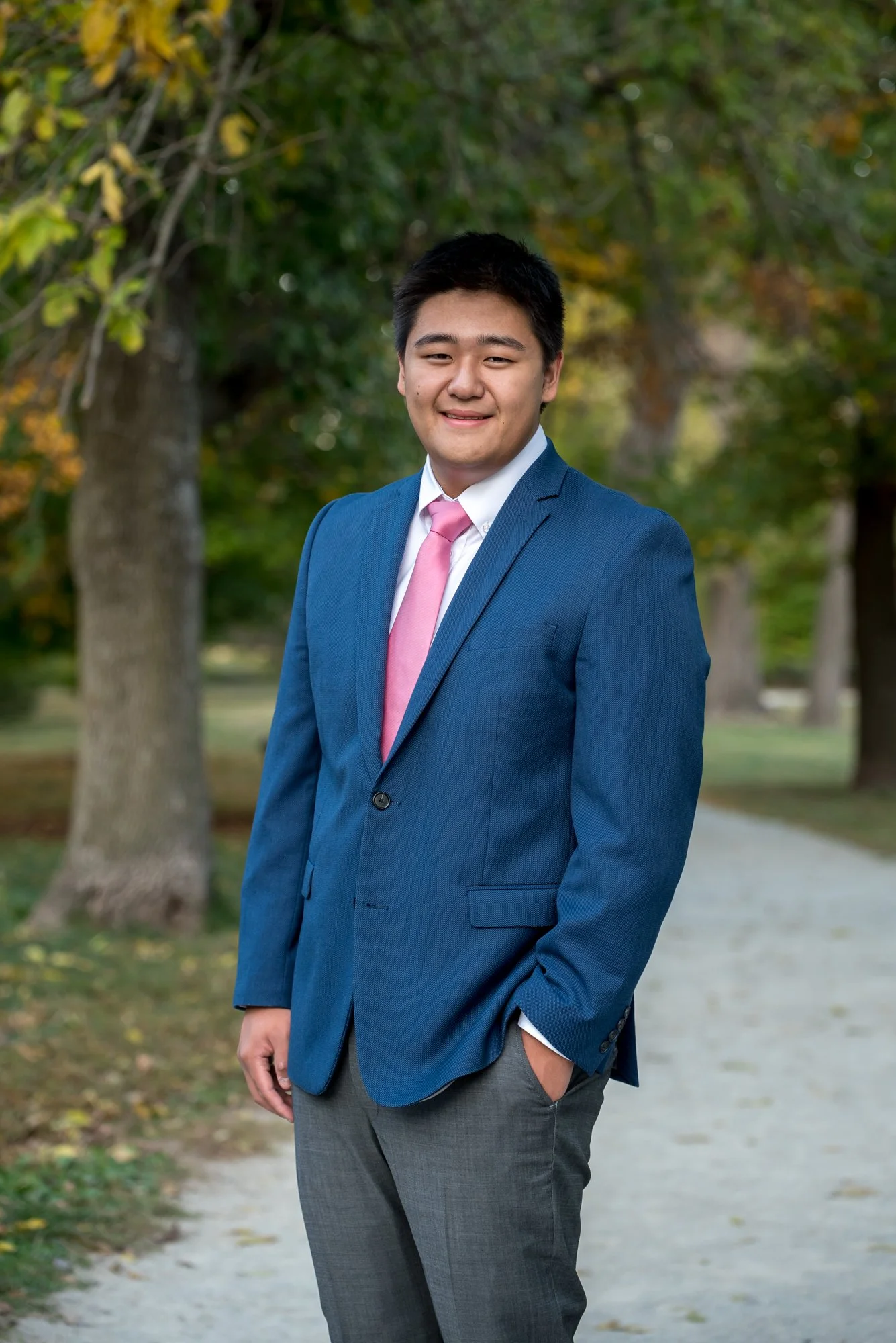 Professional-style senior picture of a boy walking along a park pathway bordered by peak fall colors, wearing a navy blue blazer and gray dress pants with one hand in his pocket taken by St. Louis senior photographer.
