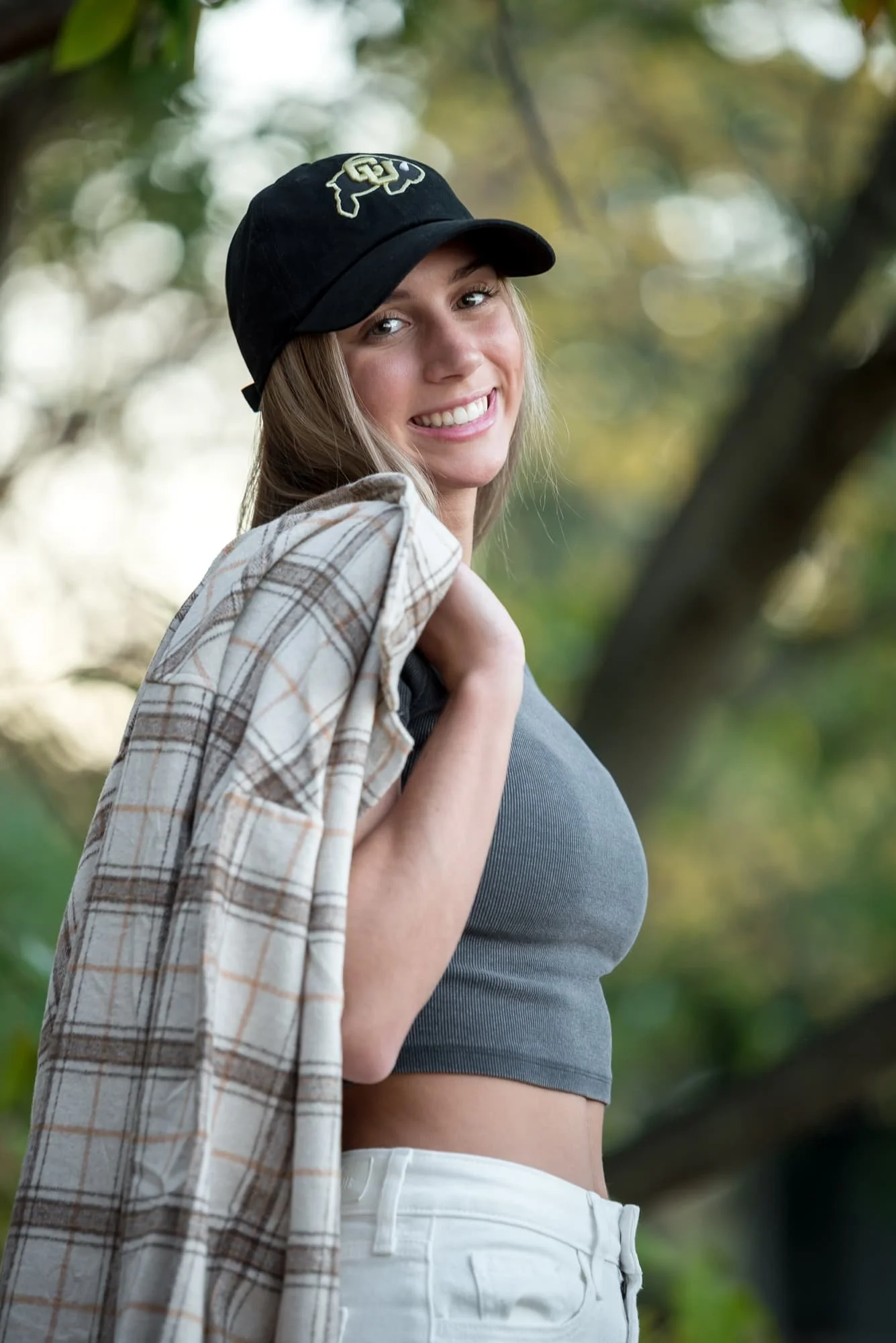 Stylish close-up senior portrait of a girl wearing a university baseball cap and gray crop top with white pants, casually throwing her tan plaid button-up shirt over her shoulder in Forest Park.