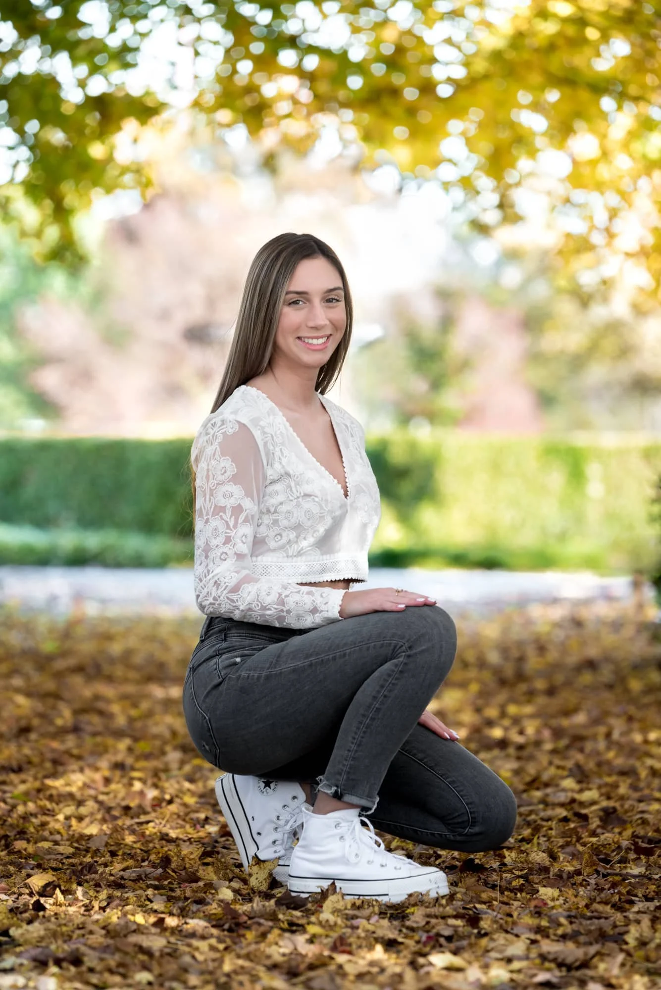 Whimsical fall senior photo taken at the Jewel Box, capturing a girl in a floral white long-sleeve crop top and black skinny jeans, crouching down under a canopy of framing autumn trees.