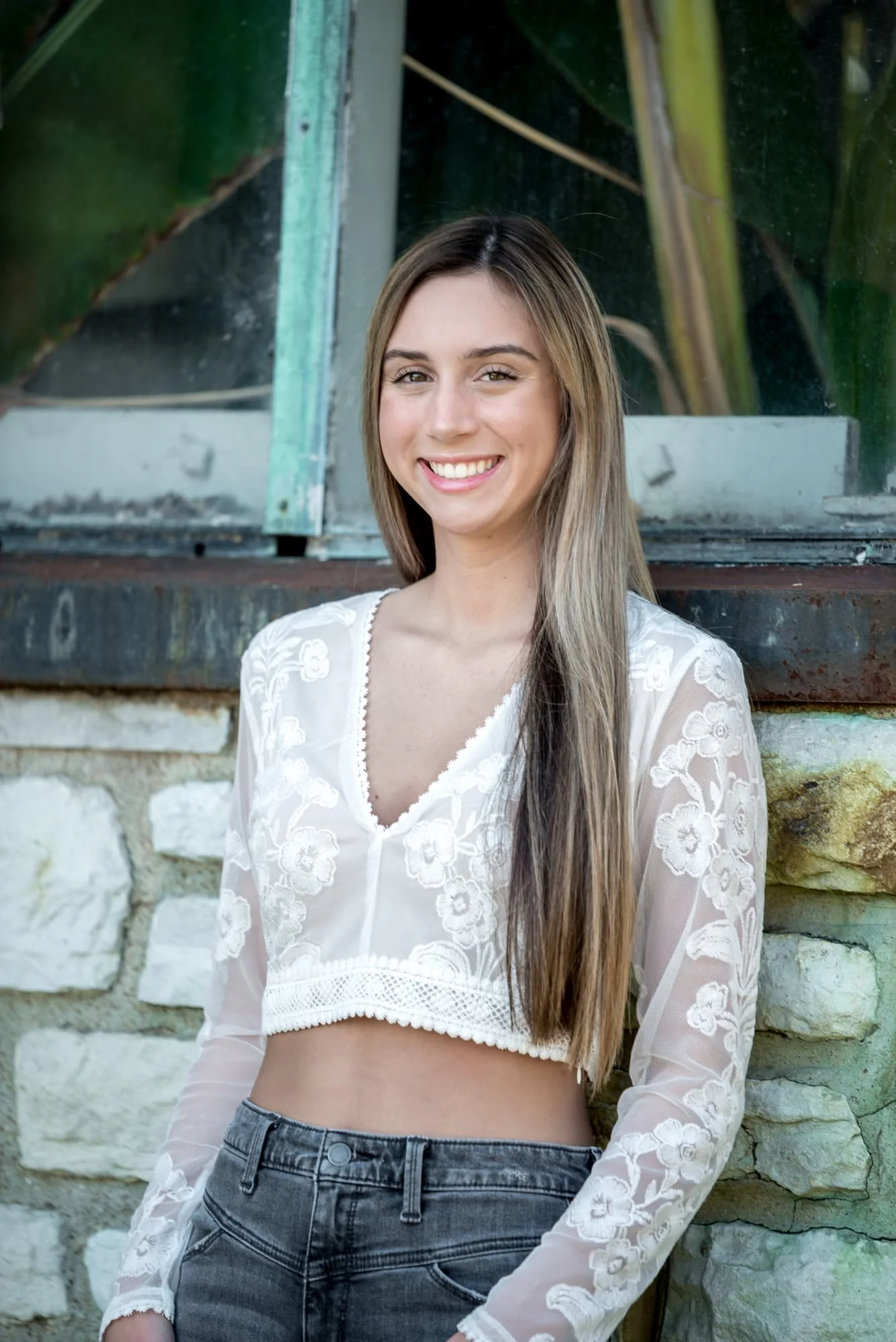 Close-up architectural senior picture showing a girl in a white long-sleeve crop top and engraved floral jeans, leaning back against the iconic stone and glass structure of the Jewel Box building.