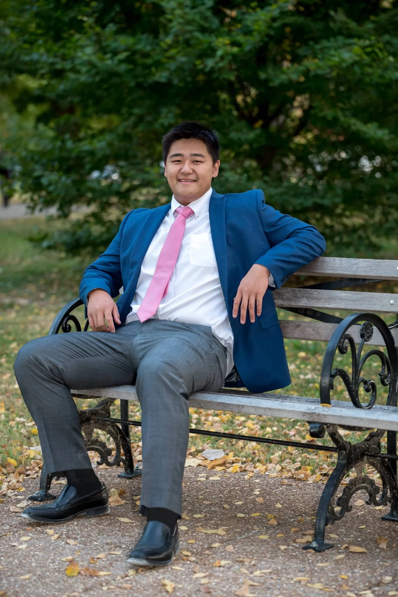 Sharp senior portrait by a St. Louis senior photographer, showing an Asian guy in a navy blue blazer, white button-up shirt, pink tie, and grey dress pants, sitting relaxed on a park bench in Forest Park.