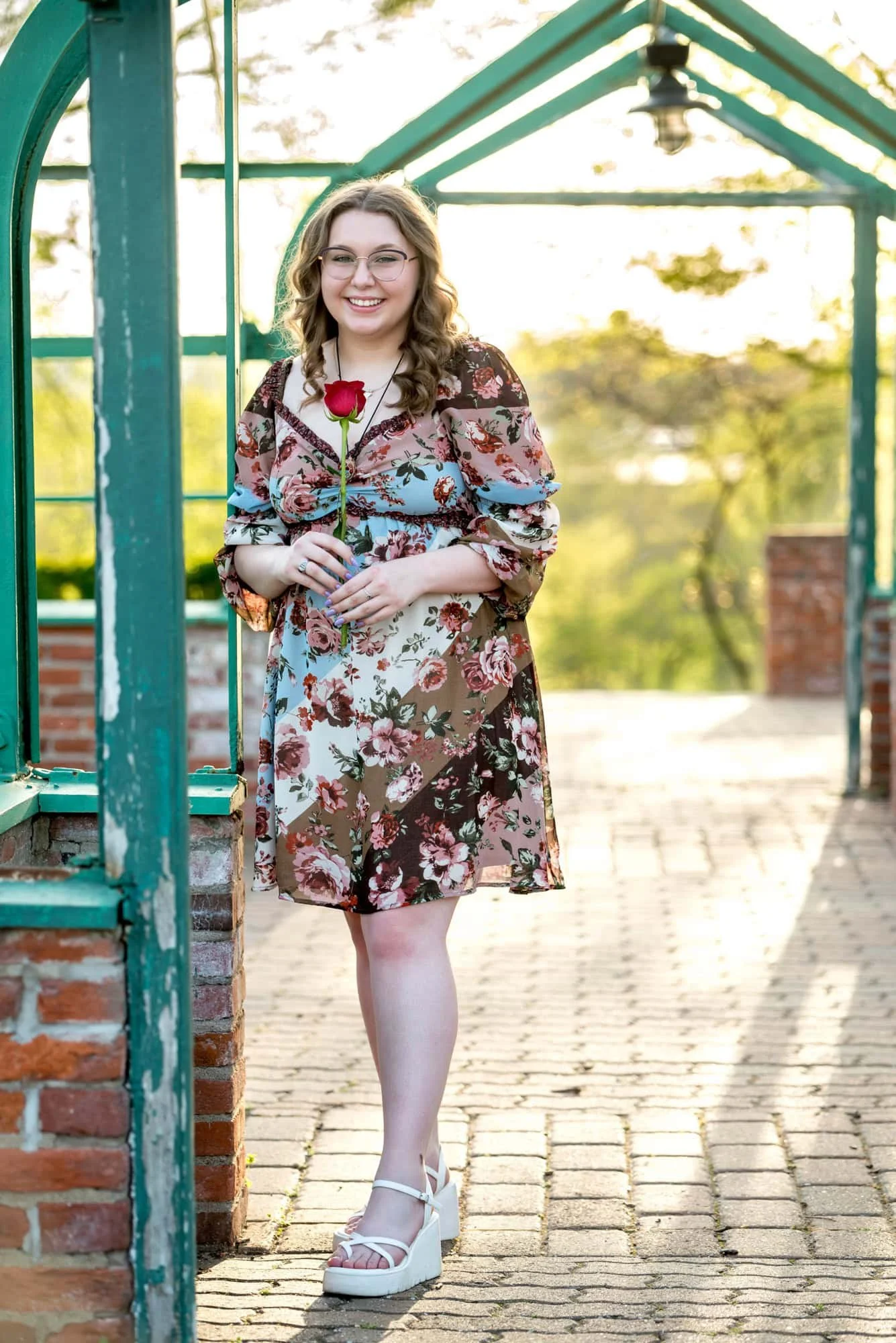 Posing outdoors at a greenhouse in Town and Country, a senior girl in a muted floral dress smiles for a sunset picture.