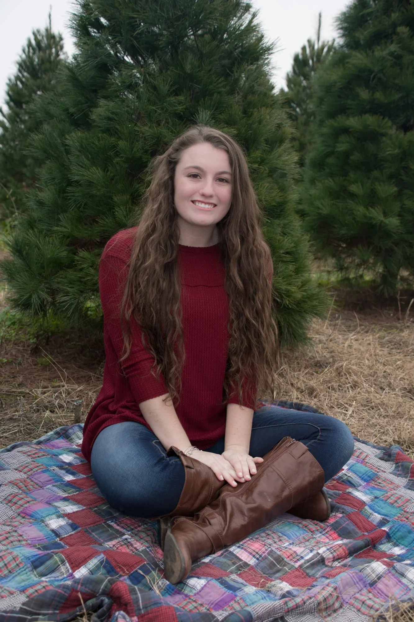 Cozy, rustic senior picture of a girl with long brown curly hair, wearing a maroon long-sleeve shirt and blue jeans while sitting on a plaid blanket nestled in the pine needles at the Eckert’s Millstadt Pine Tree Farm.