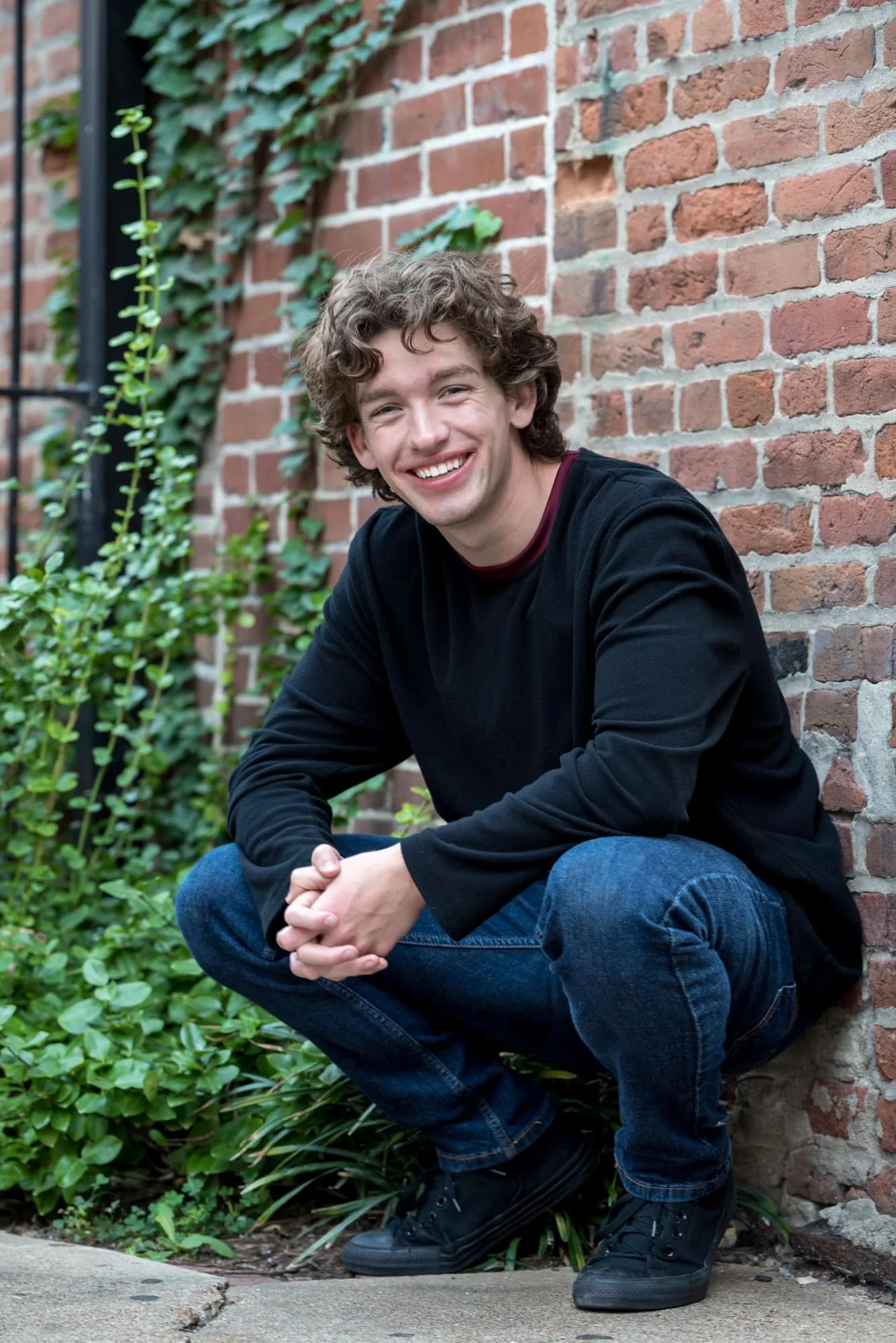 Cool, edgy senior portrait of a guy wearing a black sweater, blue jeans, and black shoes, squatting low against a rustic brick wall covered in green climbing ivy at Laclede's Landing in downtown St. Louis.