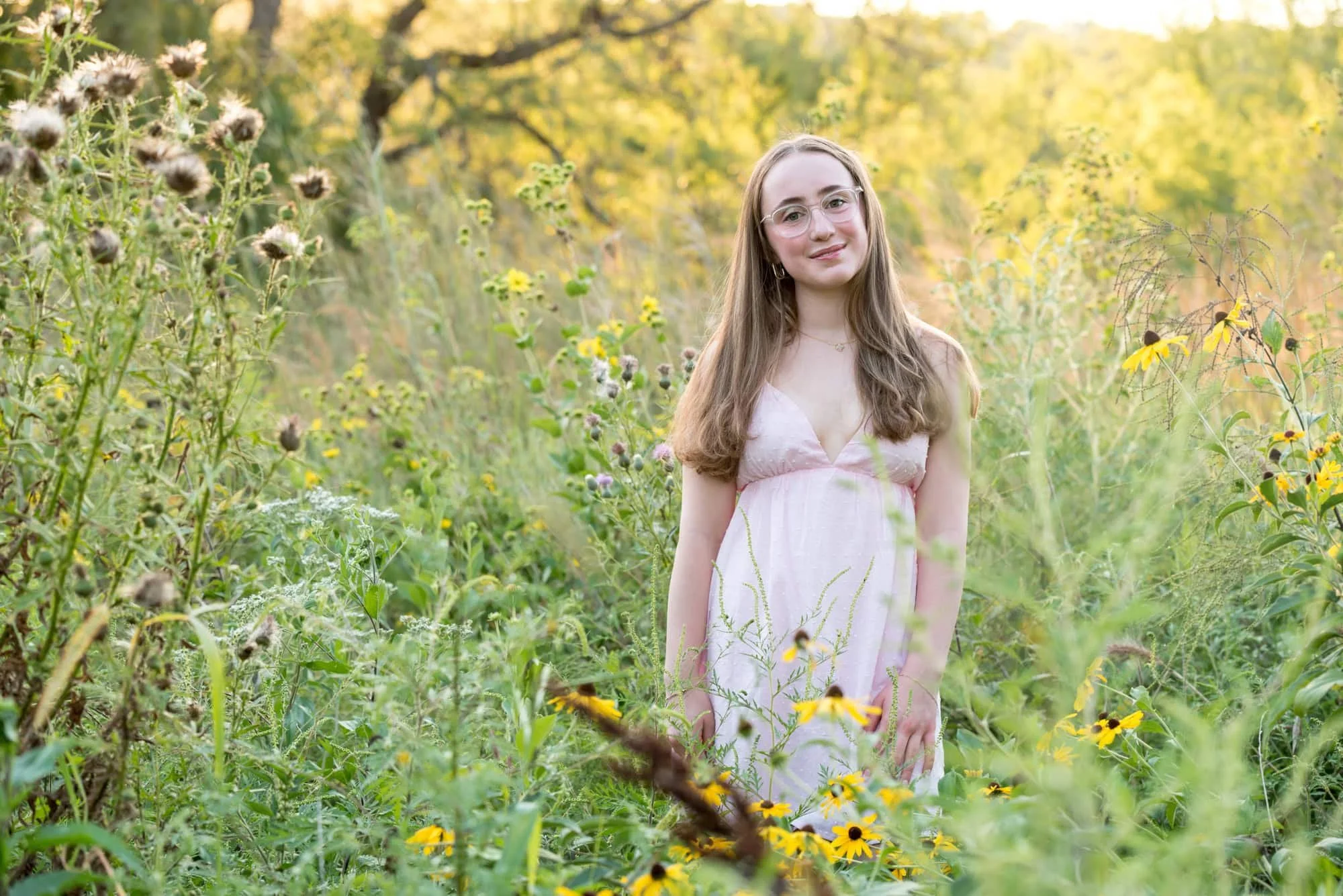 A high school senior girl wearing a pink dress and glasses standing in a wildflower field at sunset during a senior portrait session at Queeny Park.