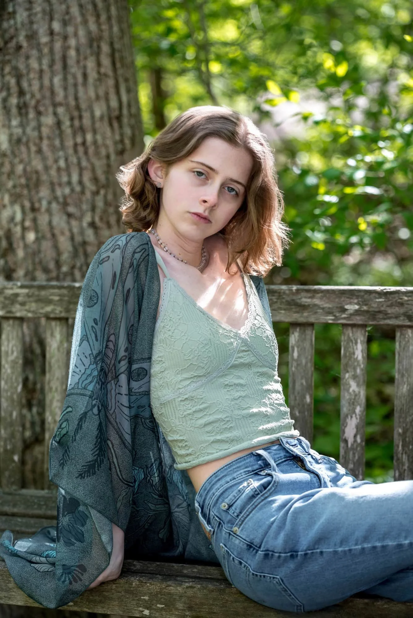 Natural light close-up photo of a senior girl leaning back against a weathered wooden garden bench, surrounded by the foliage of Gray Summit, Missouri, wearing a green lace top and a teal pattern kimono.