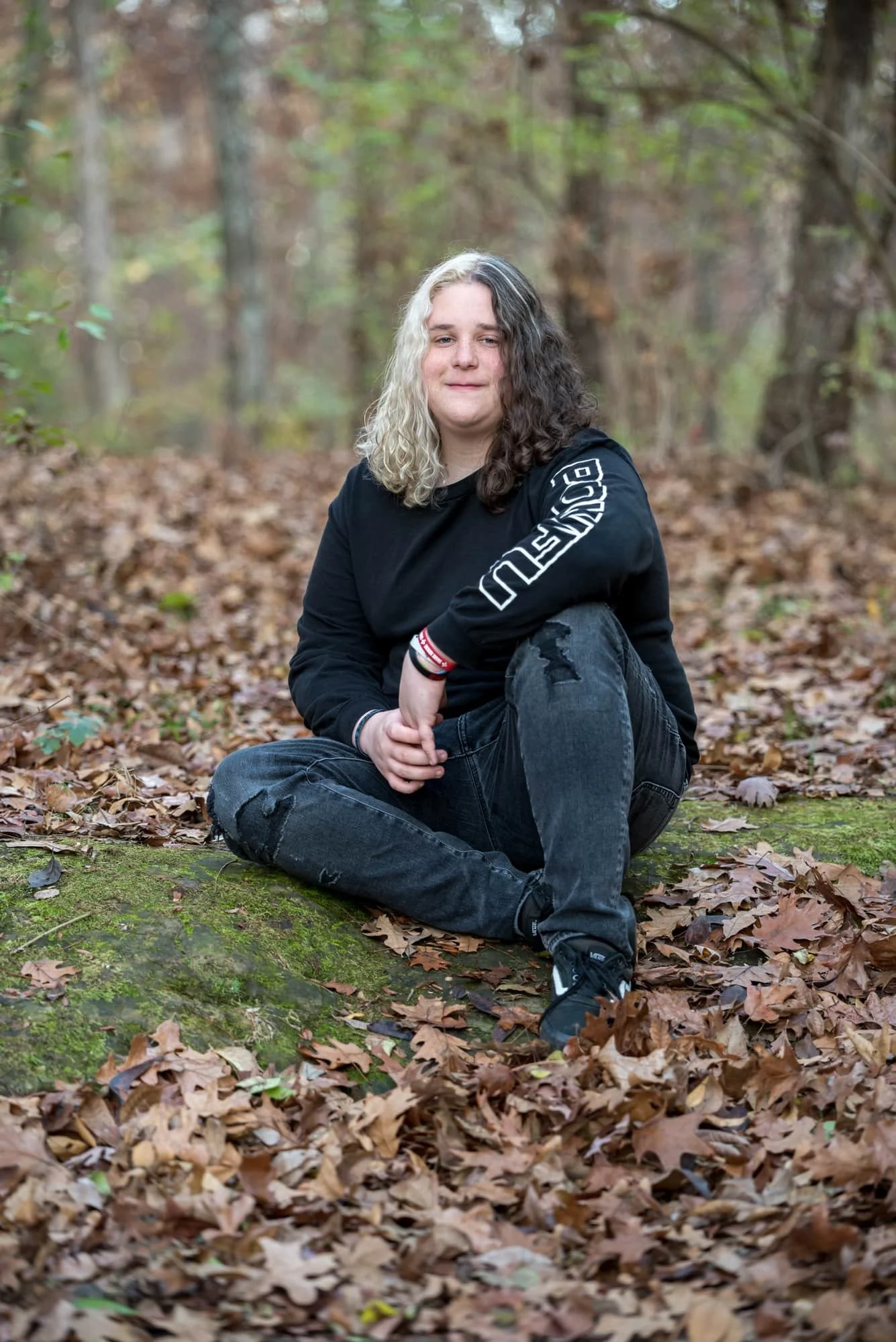 Edgy and moody senior photo of a guy with black and white hair wearing a cozy black sweater, black ripped pants, and black shoes, sitting thoughtfully on a mossy rock deep in the autumn woods.