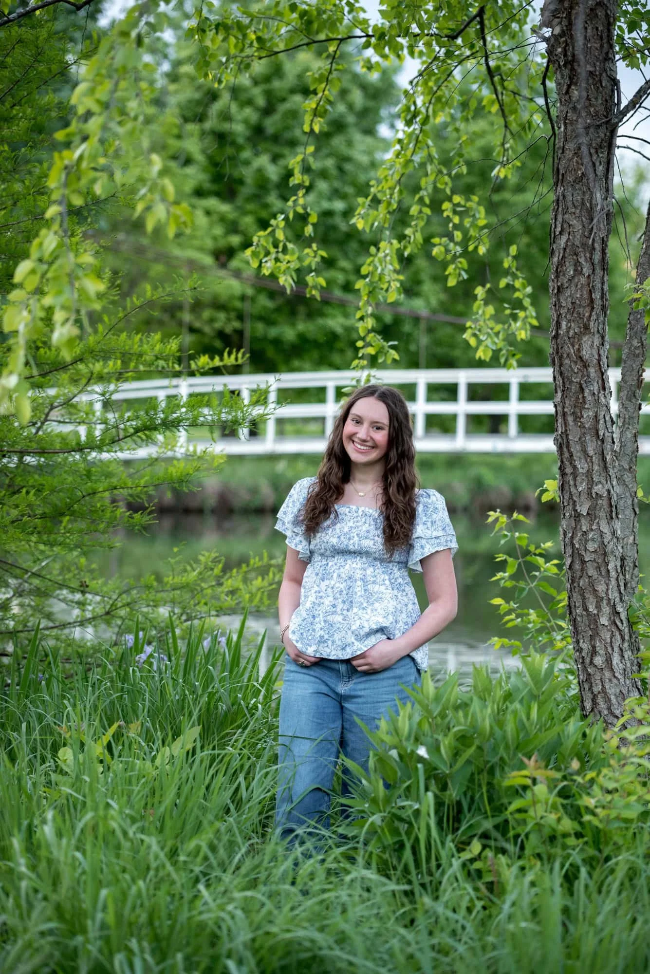 Surrounded by lush green foliage and overhanging branches in Forest Park, a senior girl in a casual blue top smiles in front of a white wooden bridge over the water.
